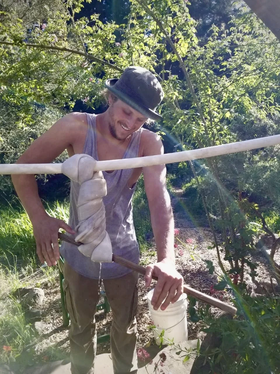 A man smiling while working outdoors surrounded by green foliage, holding a long stick with a wrapped cloth around it and a white bucket on the ground nearby.