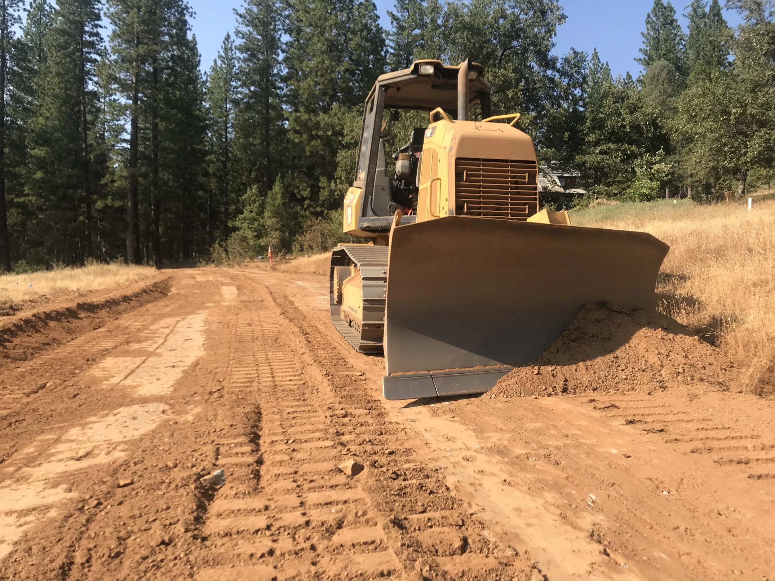 A bulldozer working on a dirt road in a forested area with trees and a house in the background.