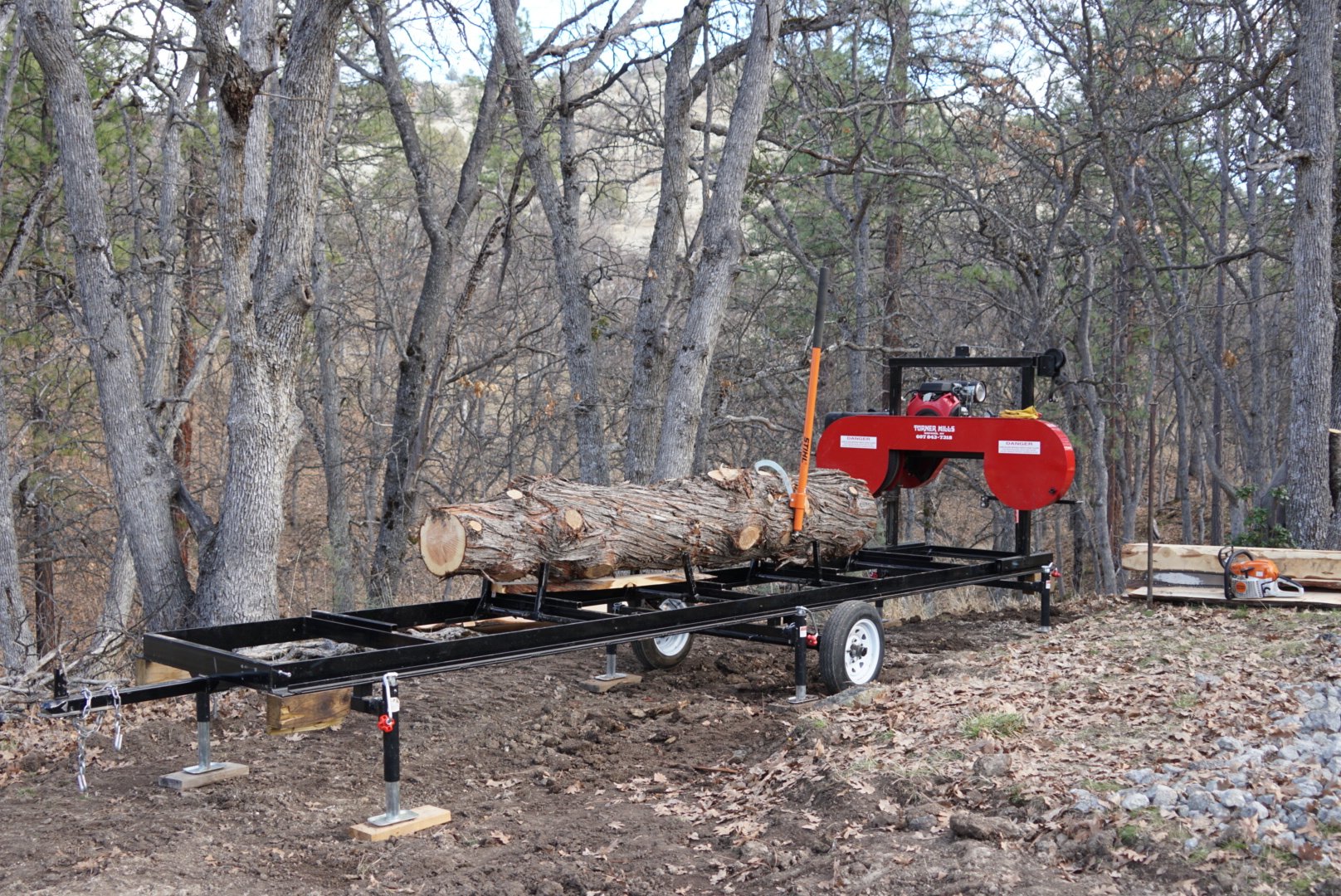 A chainsaw mill setup in a wooded outdoor area, cutting a large log on a portable framework with trees and dirt ground in the background.