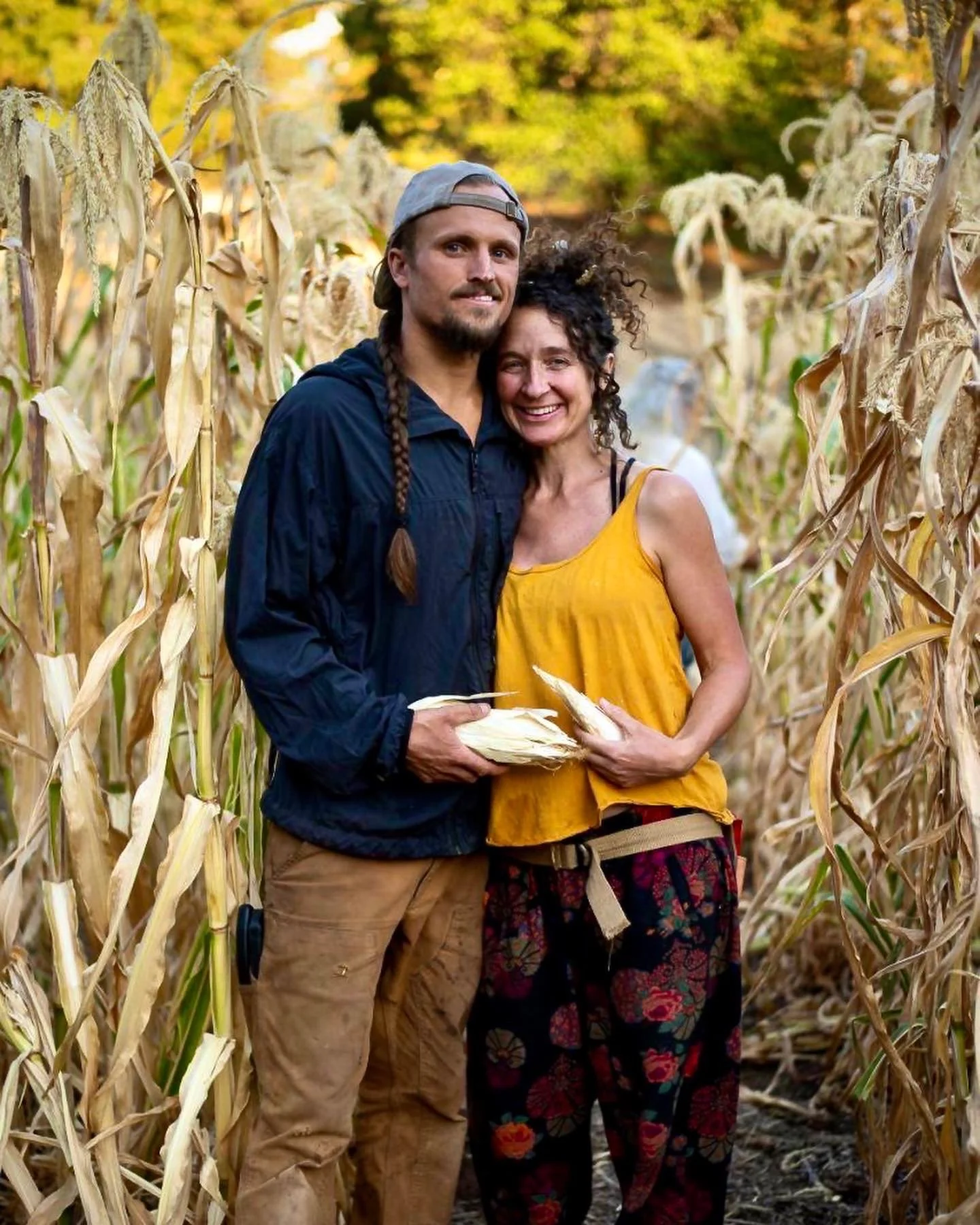A smiling couple stands in a cornfield, holding ears of corn, with autumn foliage in the background.