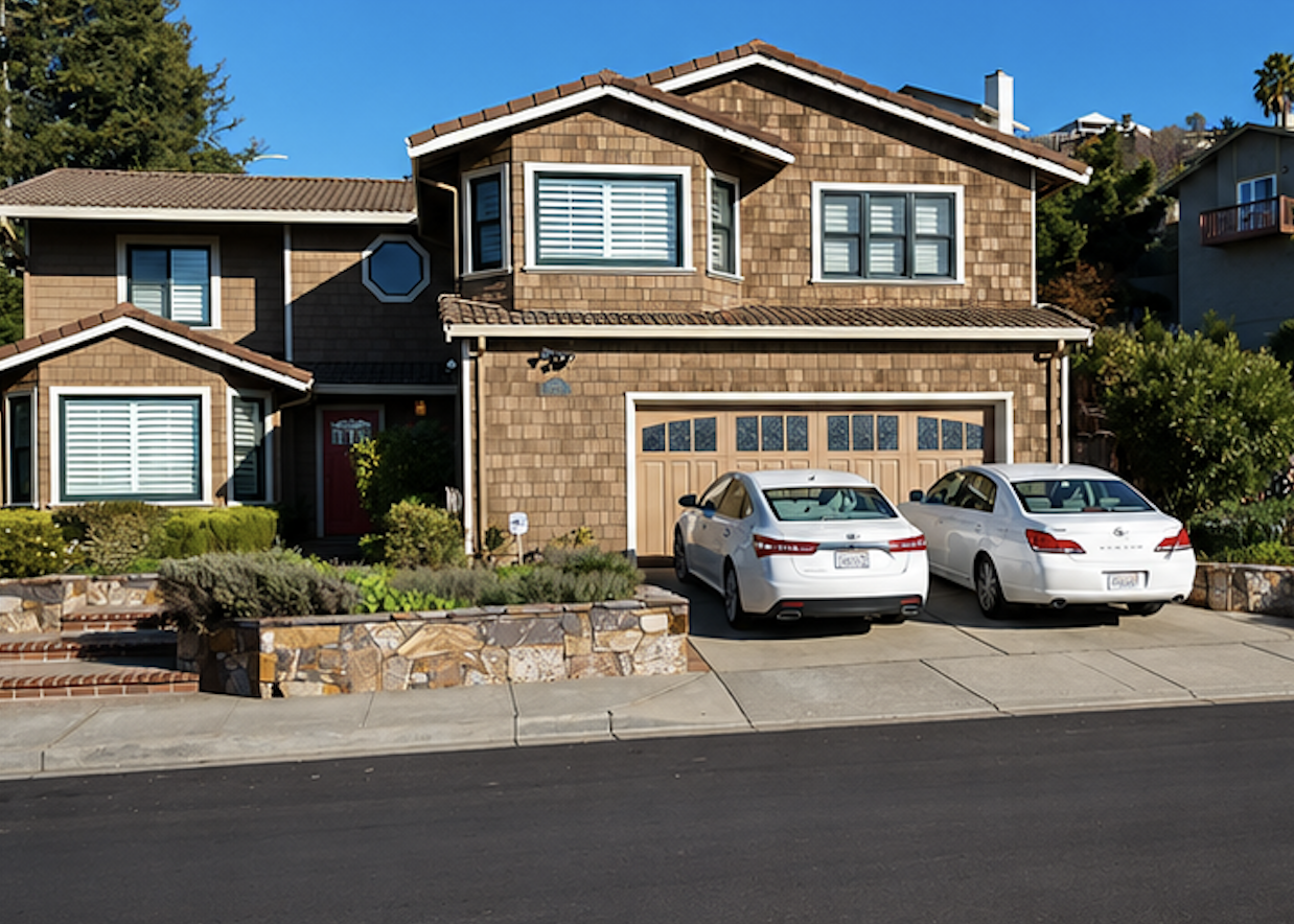 Two white cars parked in front of a two-story house with a stone facade, brown shingle siding, and a wooden garage door, in a suburban neighborhood under a clear blue sky.