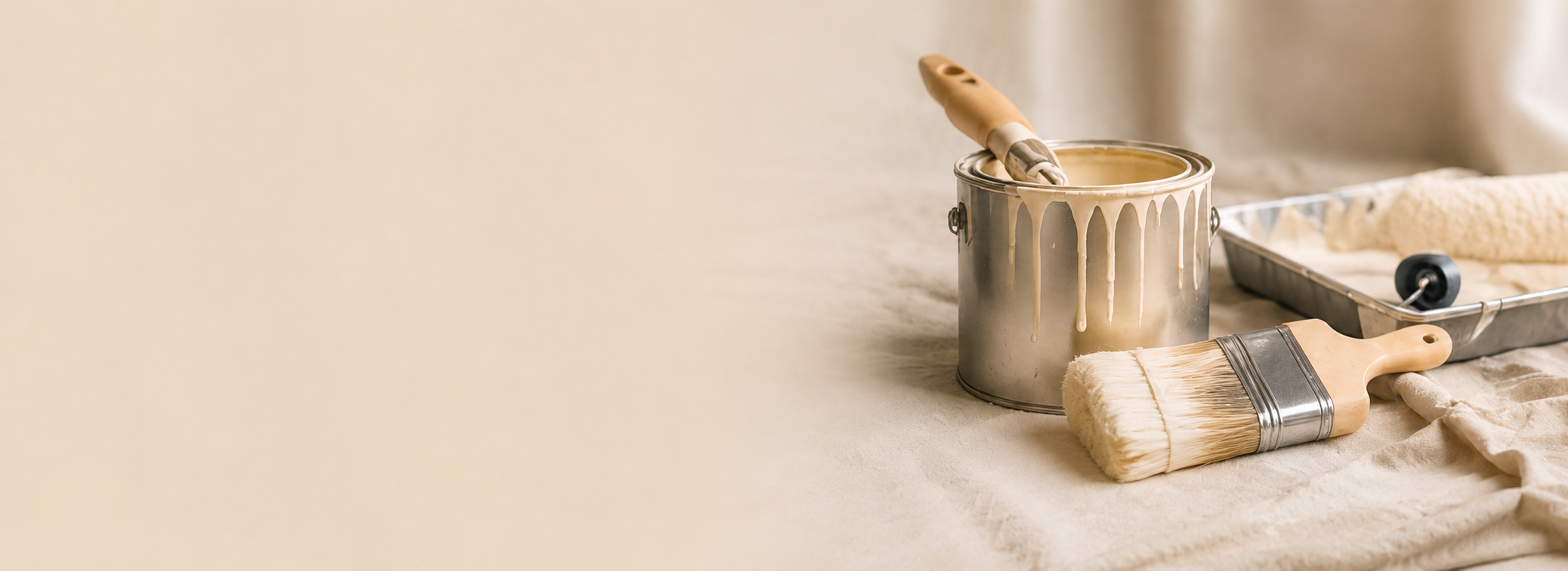 Painting supplies including a paintbrush, roller, and paint can with beige paint, arranged on a cloth-covered surface.