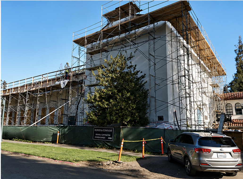 Construction site with scaffolding around a building and a tree in front, parked car, and blue sky.