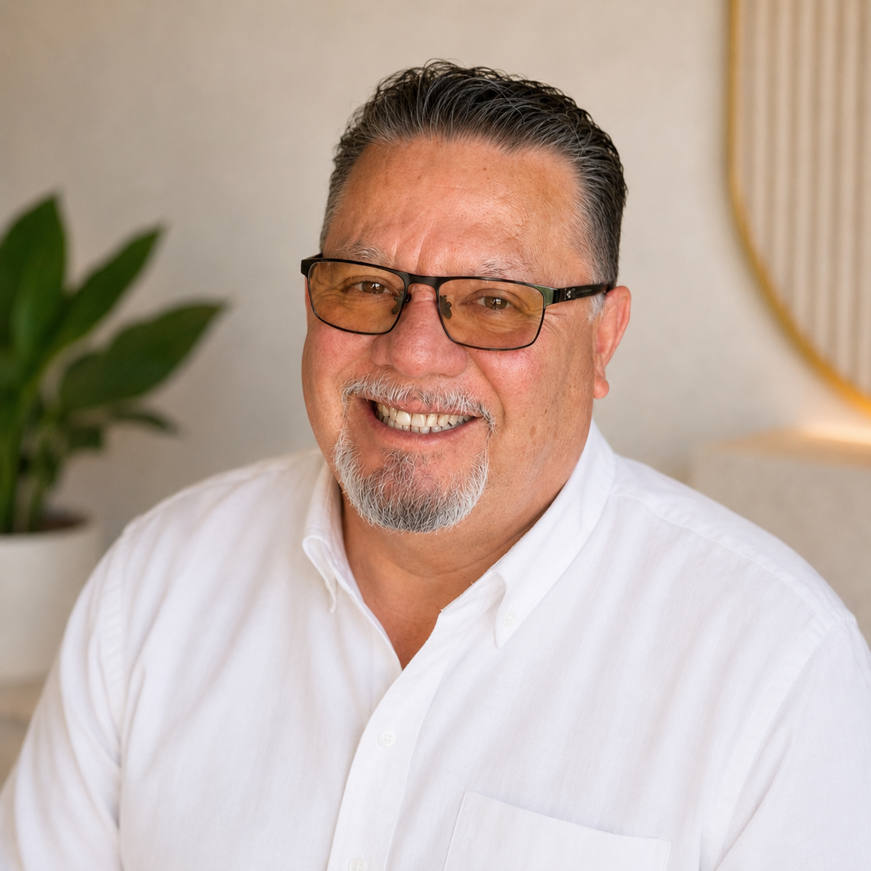 Portrait of a middle-aged man with glasses, white hair, and a beard, smiling and wearing a white dress shirt, with a plant and a mirror in the background.
