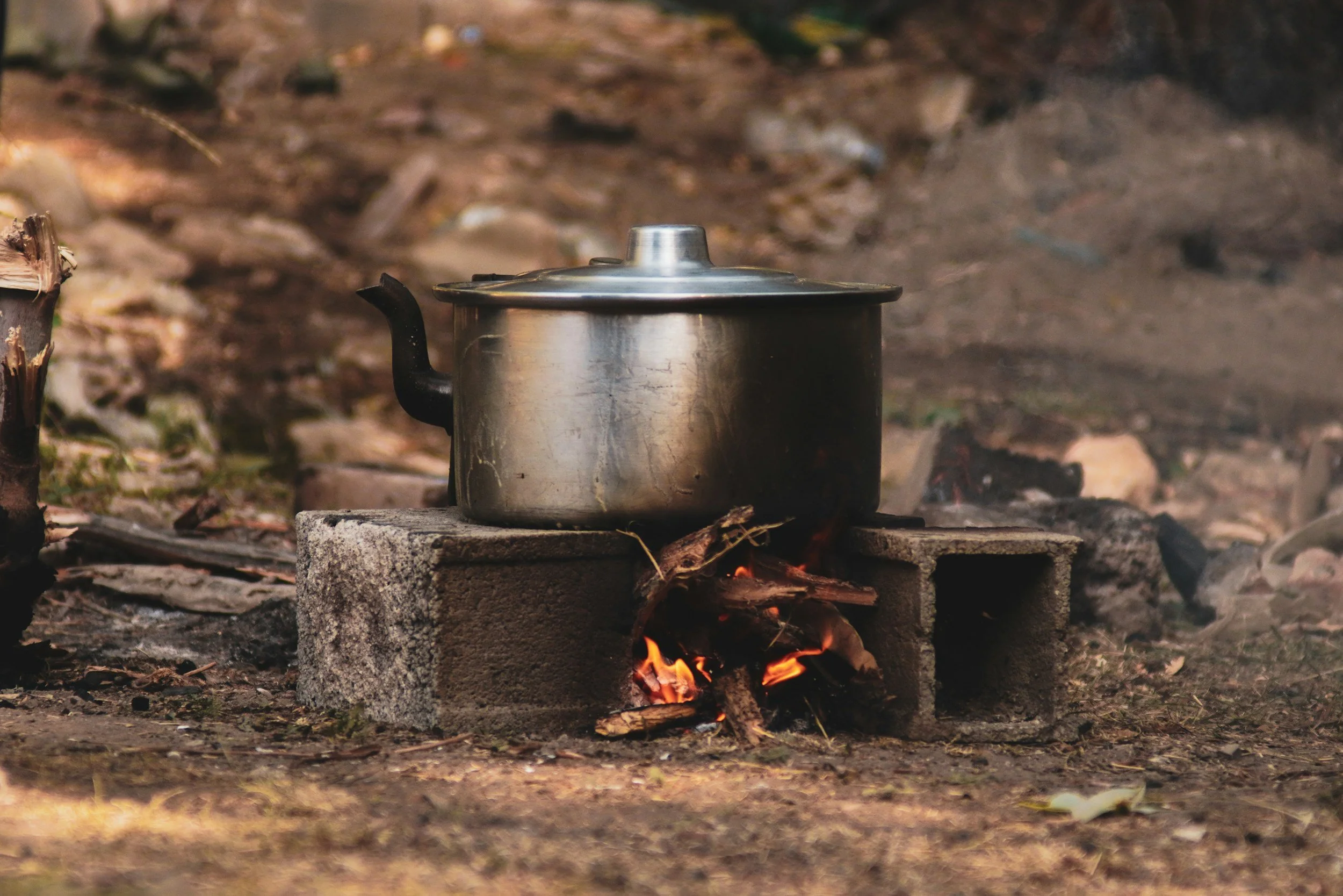 A metal kettle placed on a cinder block over a small outdoor fire, with a blurred natural background.