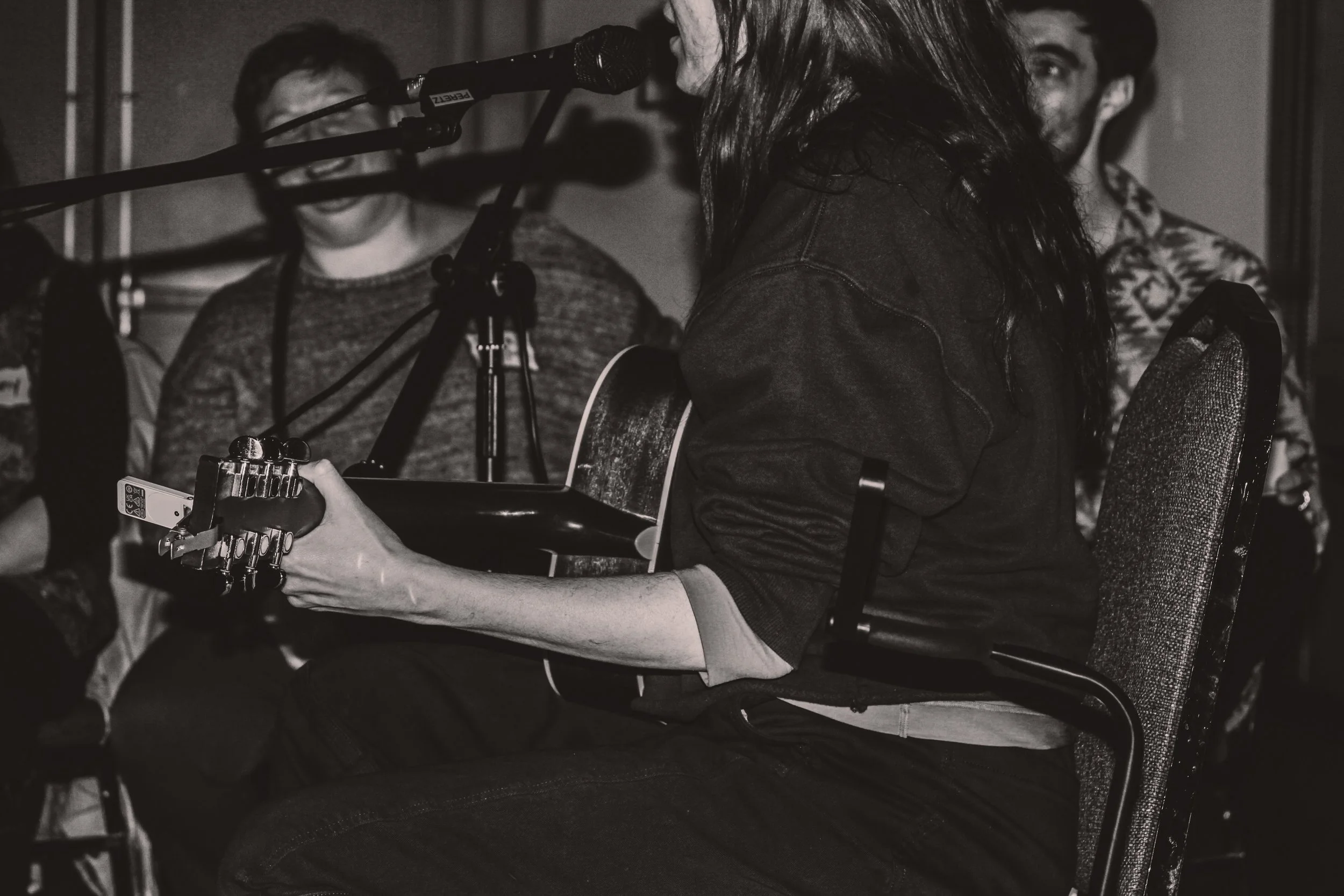 A woman playing an acoustic guitar and singing into a microphone during a live performance with two men sitting behind her.