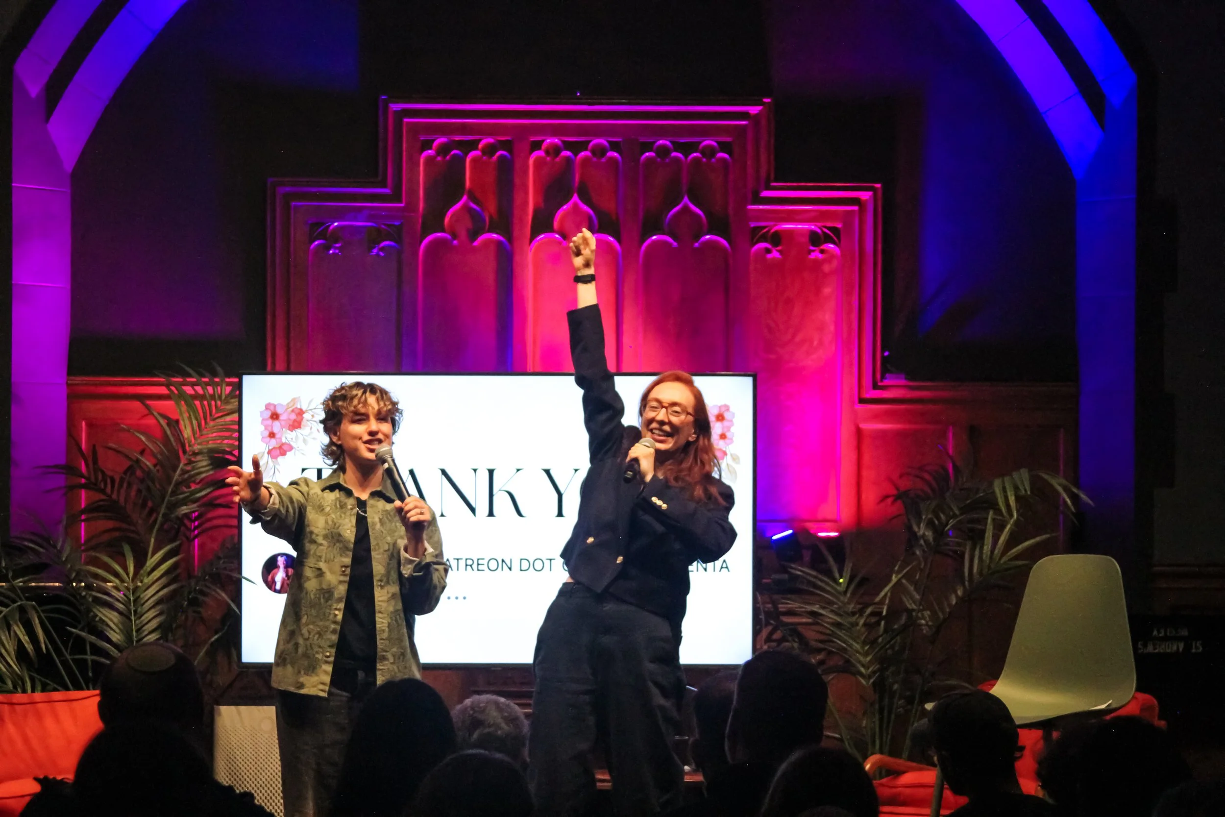 Two women speaking on stage at an event with a pink and purple lit ornate wooden background. One woman has curly hair and is holding a microphone, the other has long hair, glasses, and is raising her fist. There is a large screen behind them displayi
