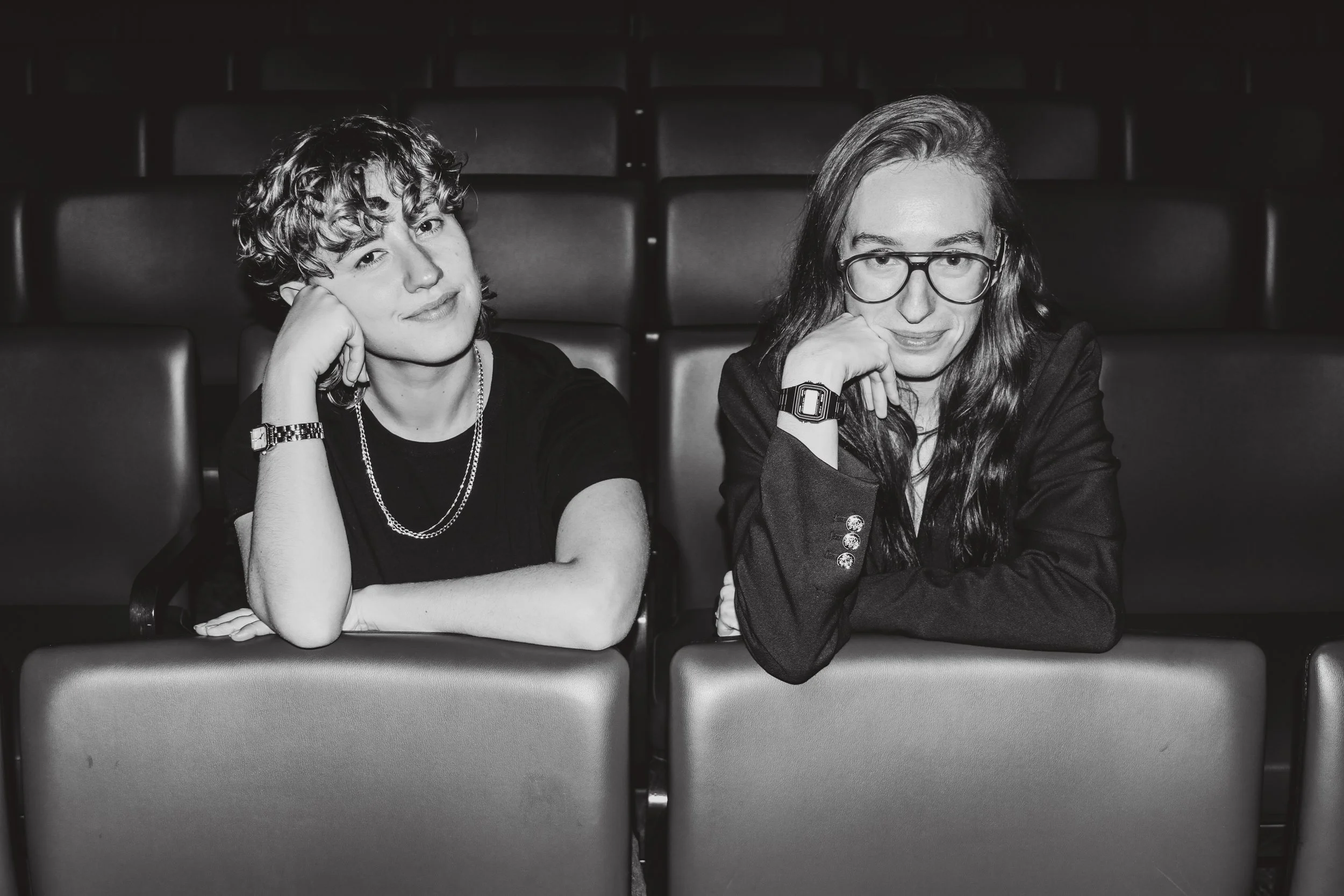 Black and white photo of two young women sitting in theater seats, resting their chins on their hands, smiling softly, with a background of empty theater seats.