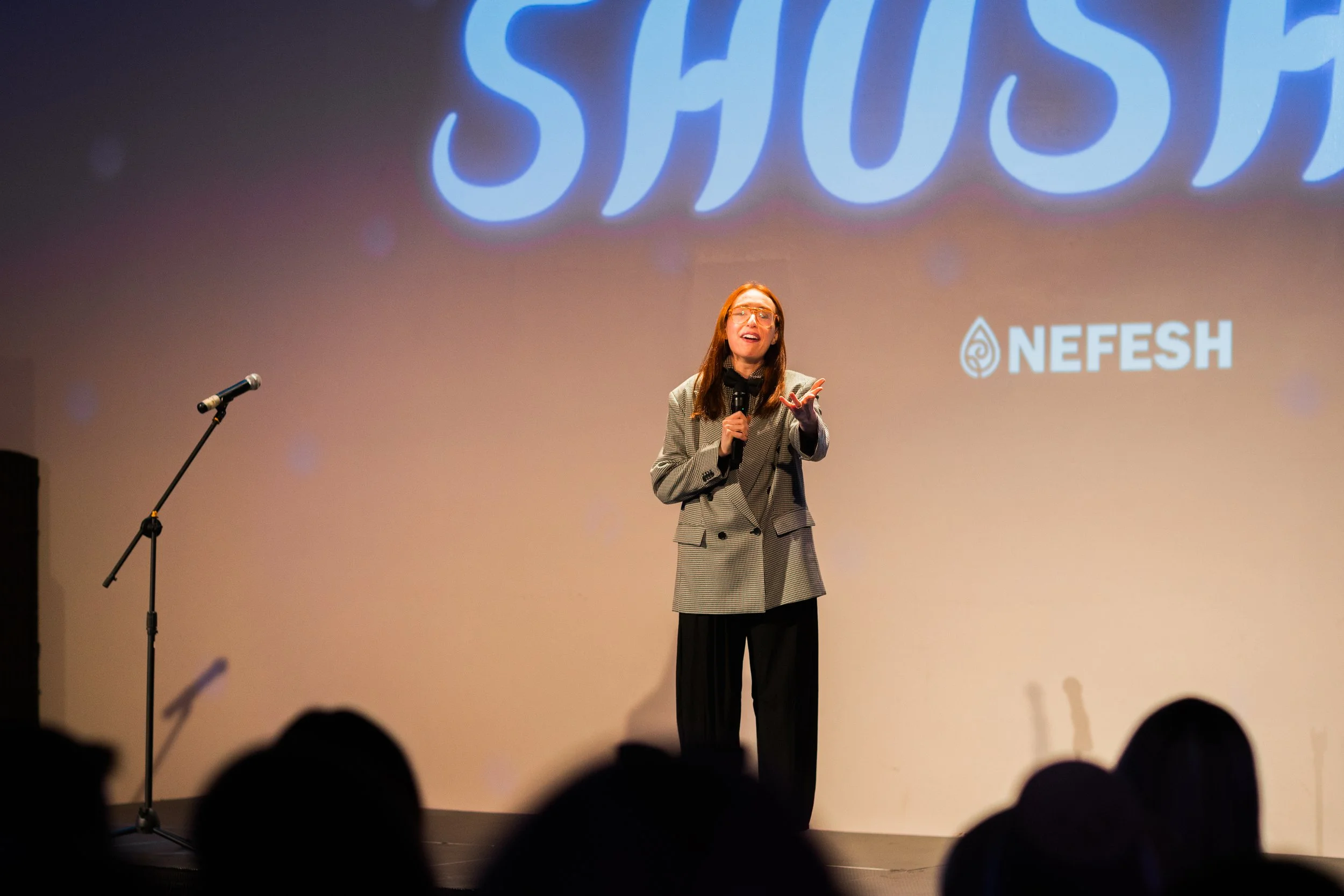 A woman speaking on stage in front of a large NEFESH logo projected on the screen behind her. She is holding a microphone and gesturing with her hand, wearing glasses and a checkered blazer.
