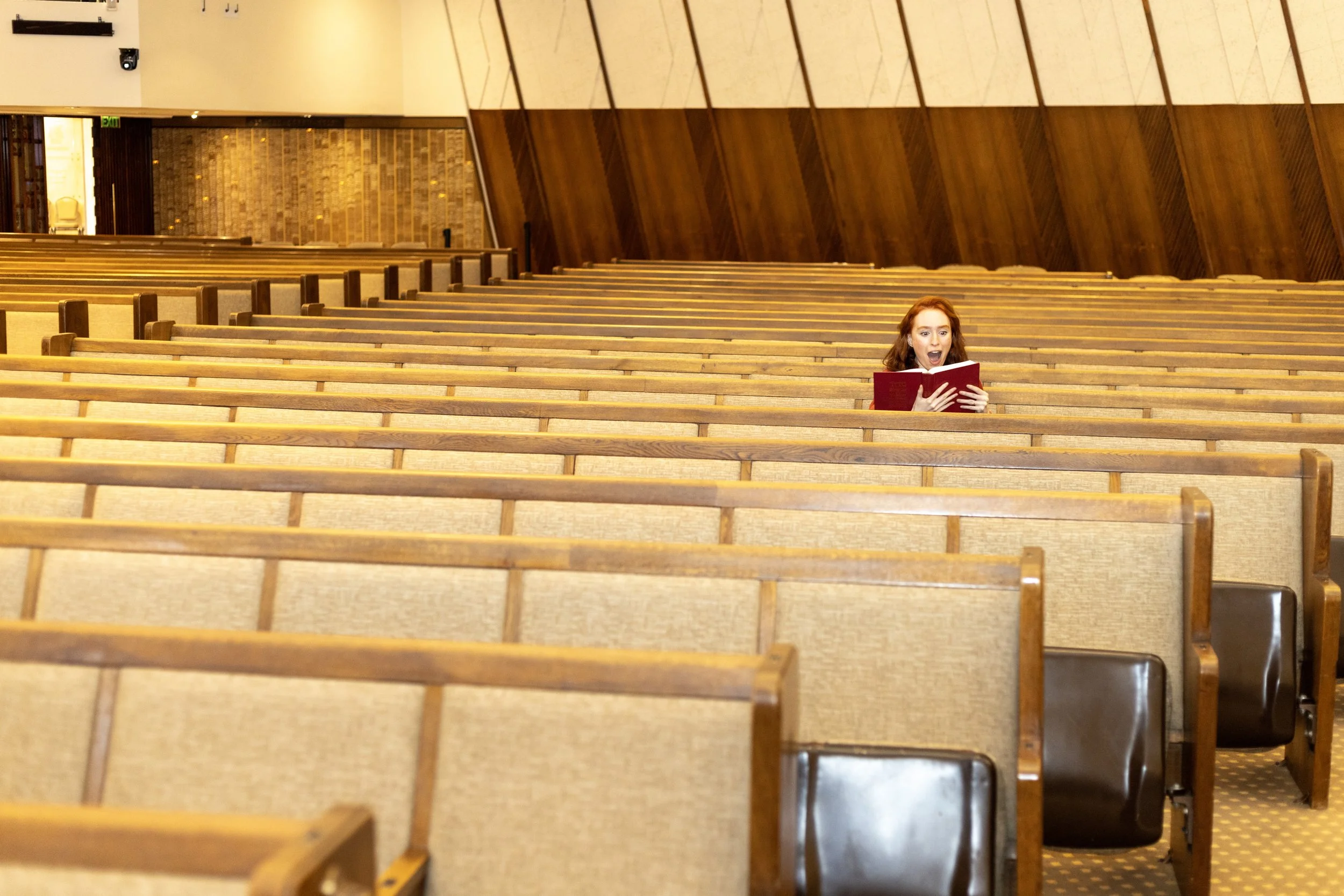 A woman with reddish hair sitting alone in a church pew, holding a red book or hymnal, reacting with surprise or shock, while the church interior is mostly empty with rows of wooden pews.