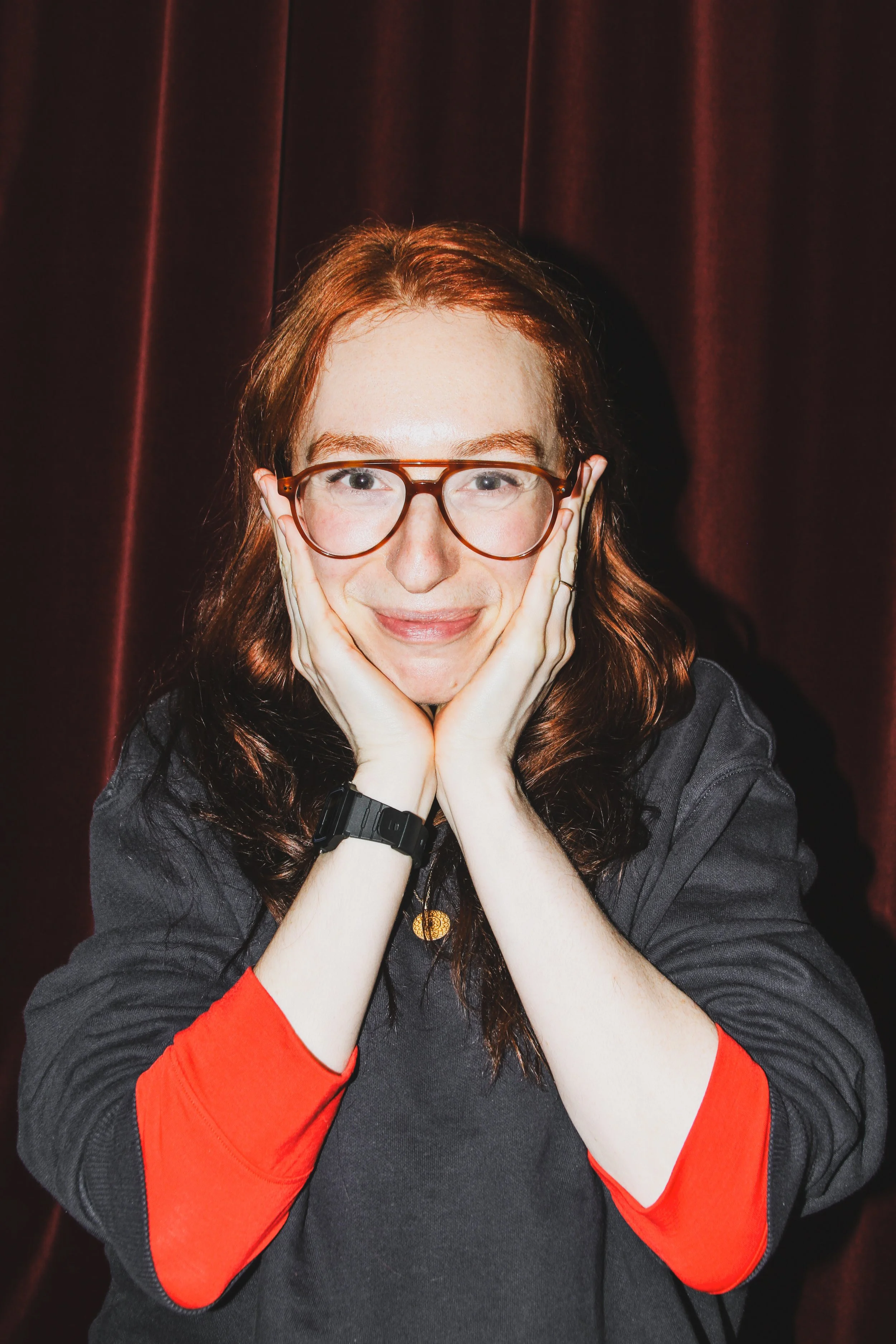 A woman with long red hair, glasses, and a black and red sweatshirt is resting her face in her hands in front of a dark red curtain.