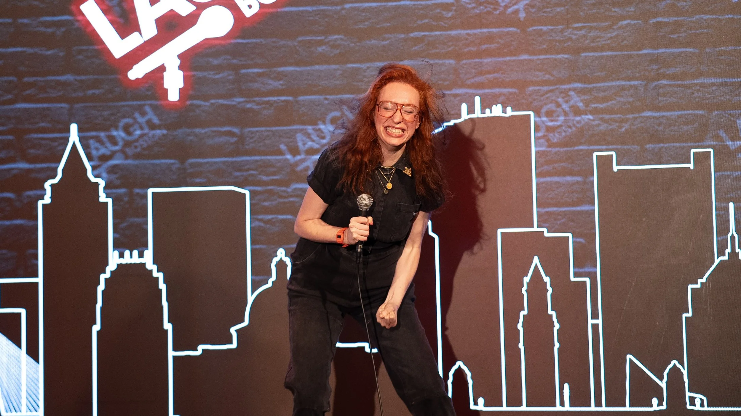 A woman with red hair, glasses, and a black outfit is holding a microphone on stage. She appears to be mid-sentence or in a moment of expression, with a backdrop of city skyline outlines and the word 'LAB' in neon above her.