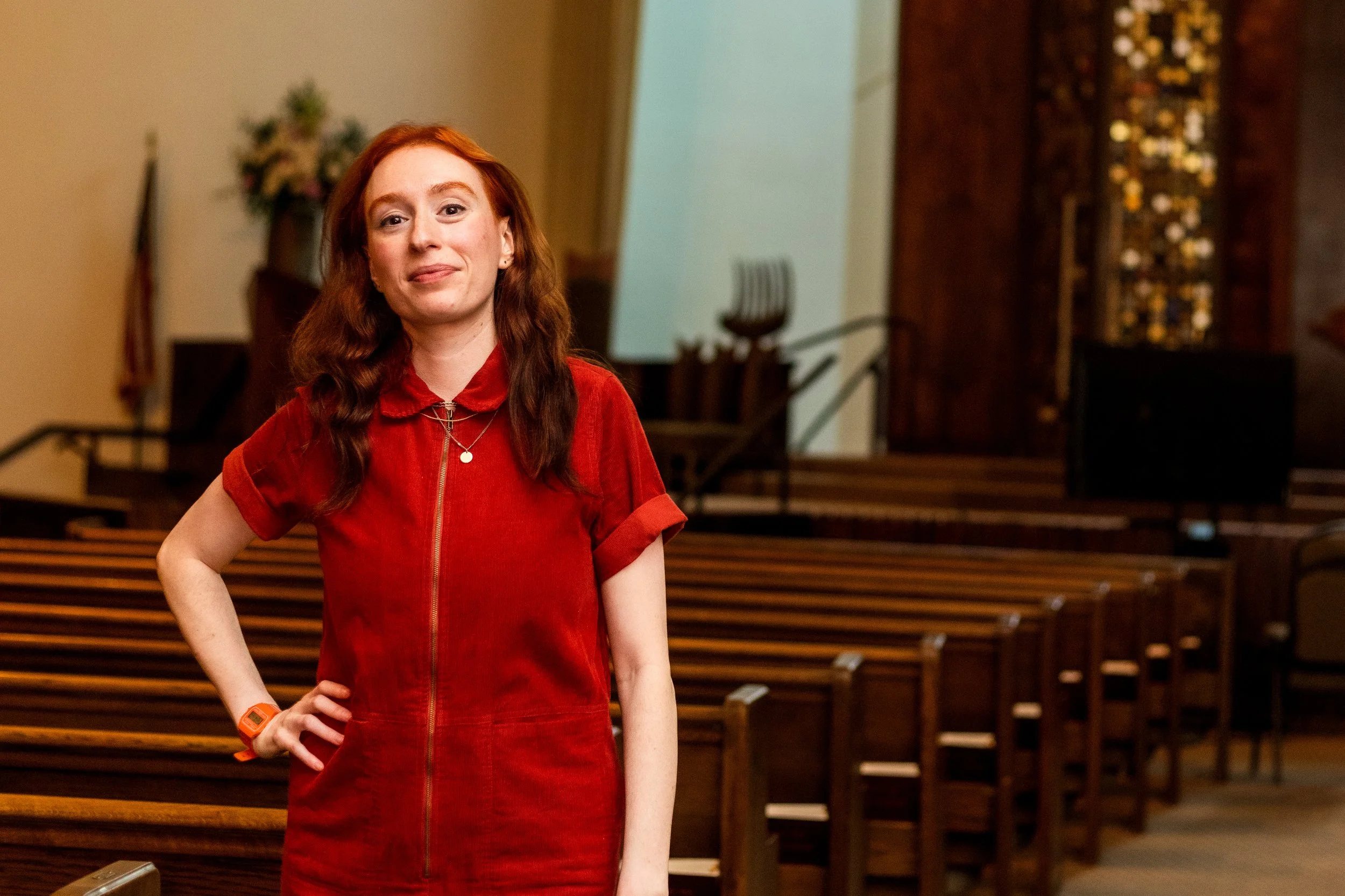 A red-haired woman in a red zip-up dress standing in an empty church with wooden pews and a floral arrangement in the background.