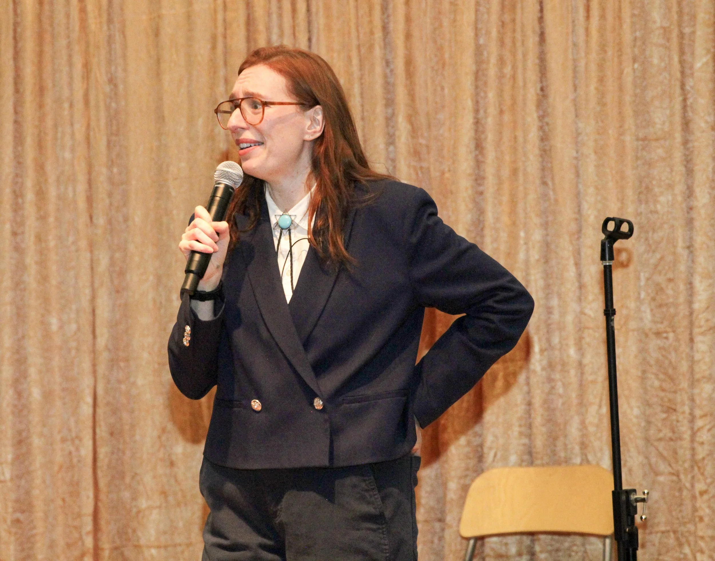 A woman with long red hair, wearing glasses, a white shirt, and a dark blazer, holding a microphone and speaking in front of a wooden paneled wall.