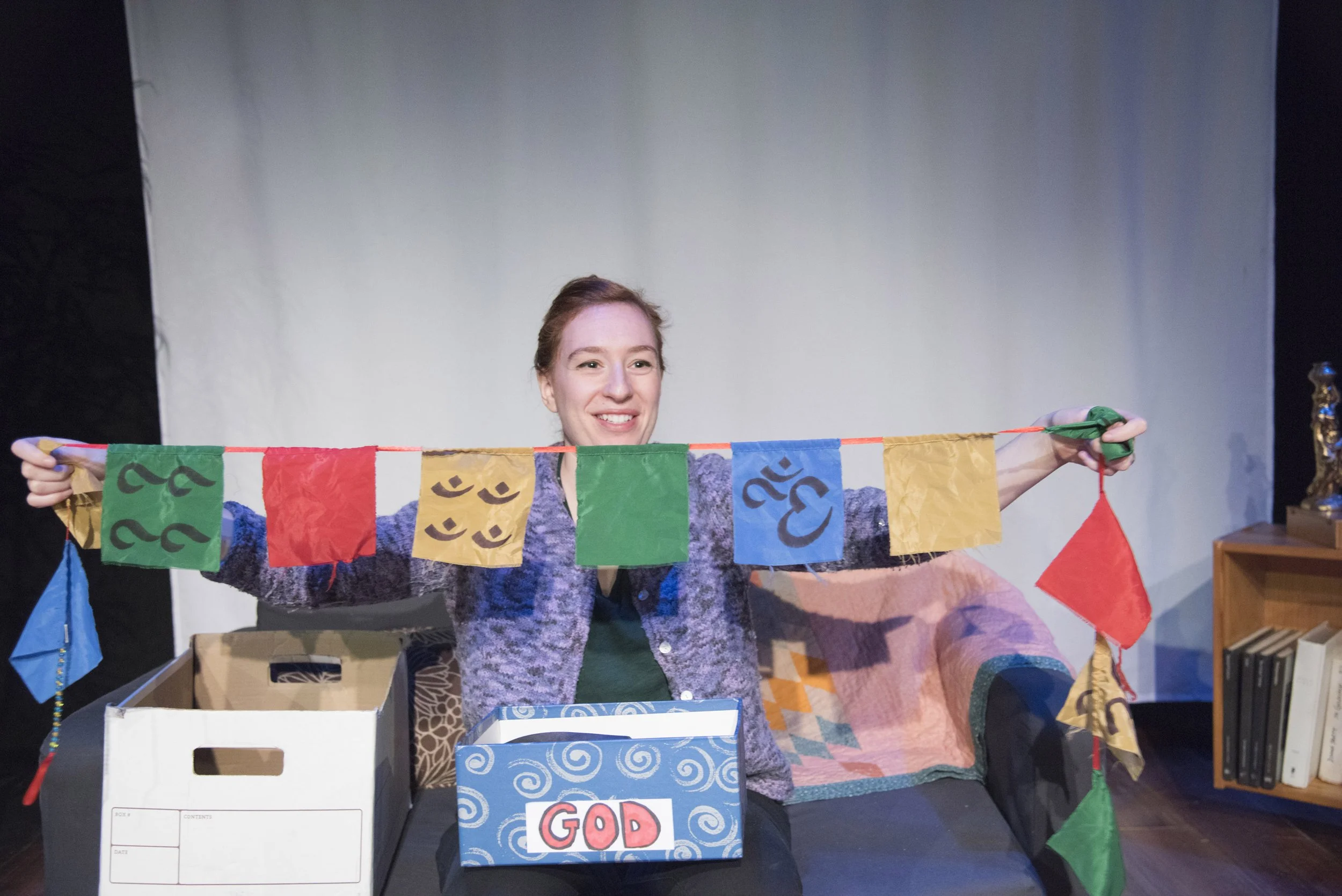 A woman with red hair holding a string of colorful prayer flags, standing behind a cardboard box with a sign that reads "GOD." There are more prayer flags and a bookshelf with books in the background.