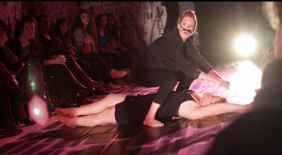 A woman in black with a fake mustache performs a dramatic act on a man lying on the floor, with an audience observing in a darkened room with stage lighting.