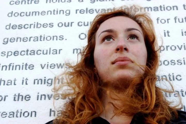 A young woman with red hair and a thoughtful expression looking upwards against a background of text on a white wall.