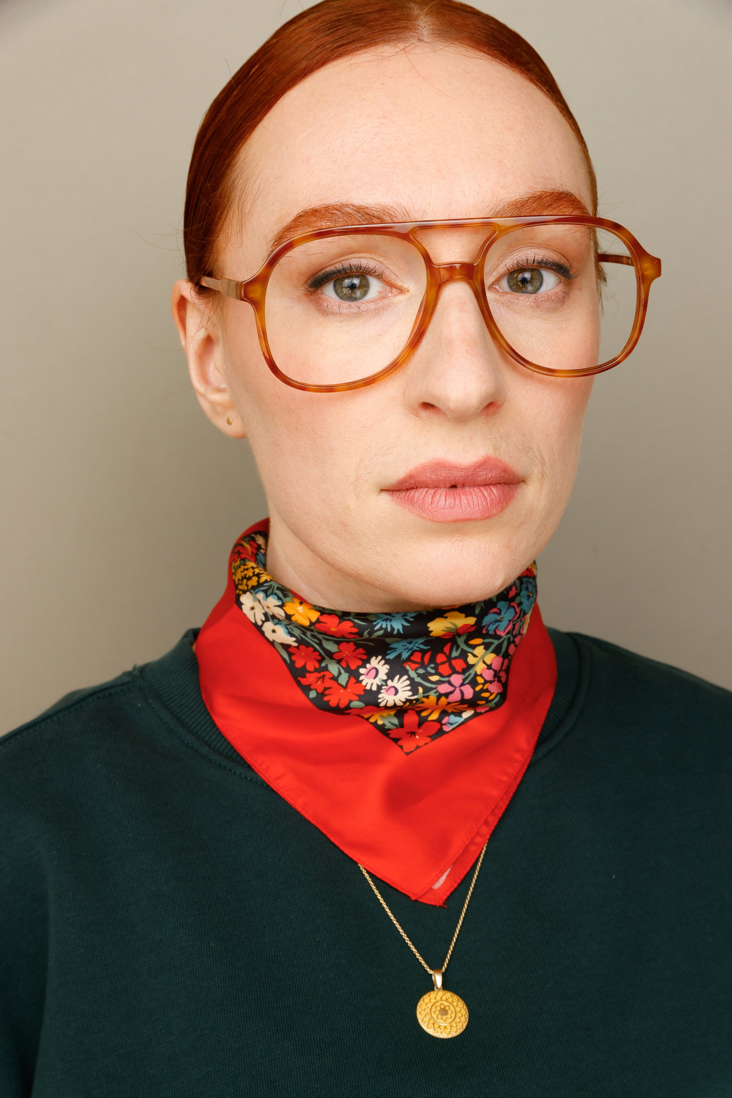 A woman with red hair wearing large cat-eye glasses, a floral scarf, a red bandana, a dark green sweater, and a gold necklace with a round pendant.