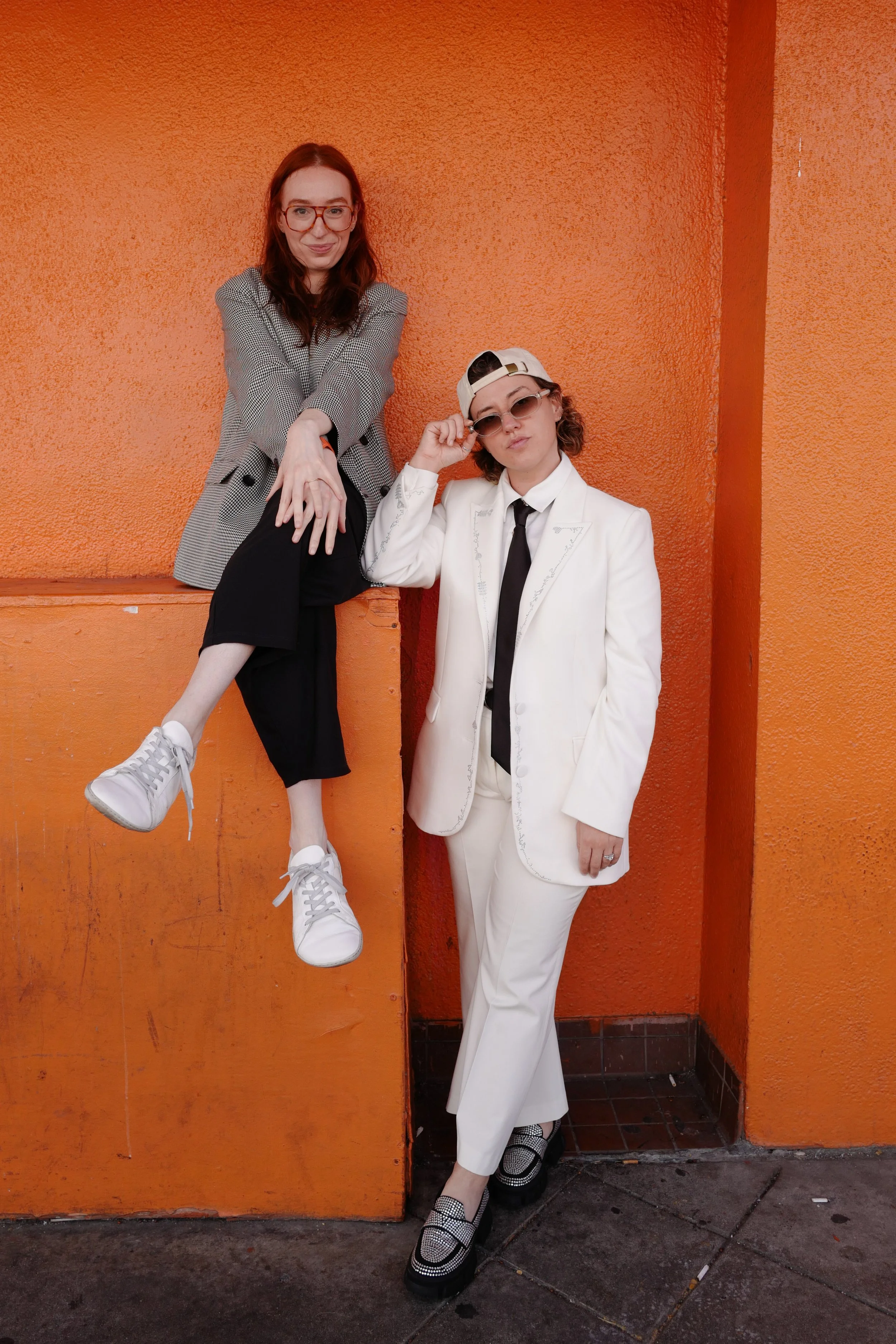 Two women posing against an orange wall, one standing and one sitting on a ledge. The woman on the left has red hair, glasses, and is wearing a checkered blazer and black pants. The woman on the right has curly hair, sunglasses, a white suit, and a c