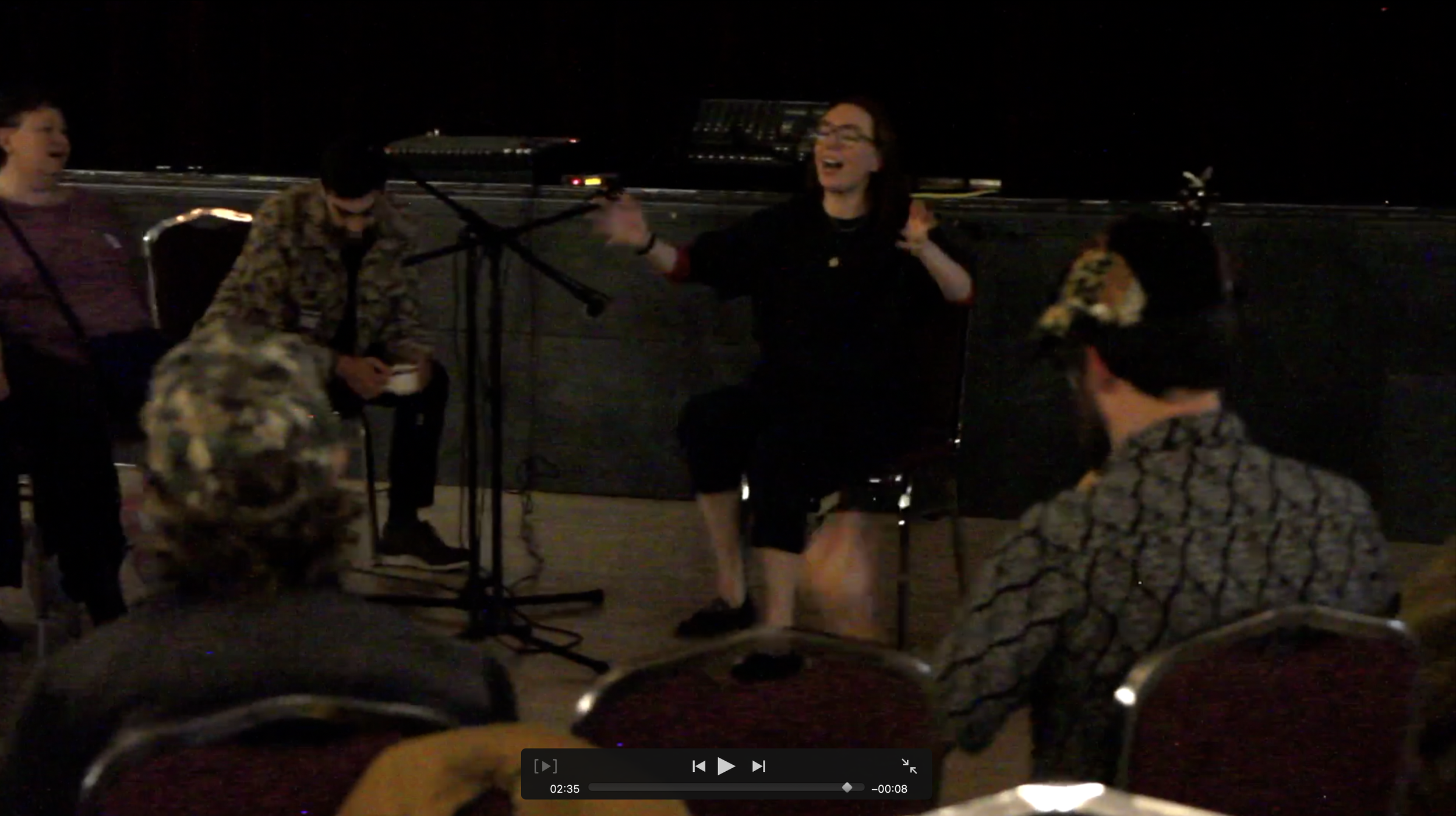 A group of people are sitting around outside at night, engaging in conversation and laughter. One woman is standing and gesturing with her hands, while others are seated nearby. There are music stands in front of them, suggesting a musical or performance setting.