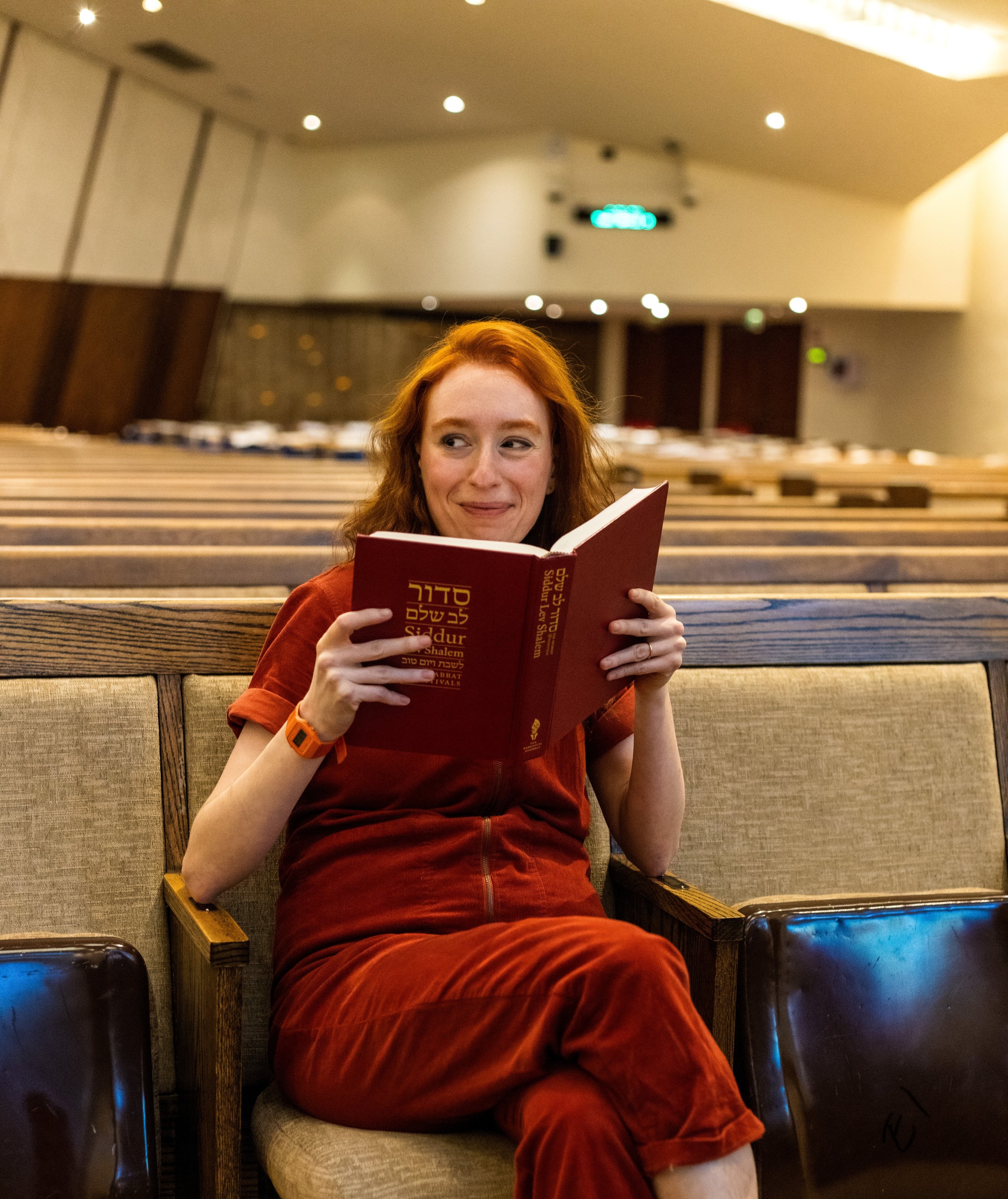 A woman with red hair wearing a red top and pants, sitting in an auditorium, smiling and reading a red book.