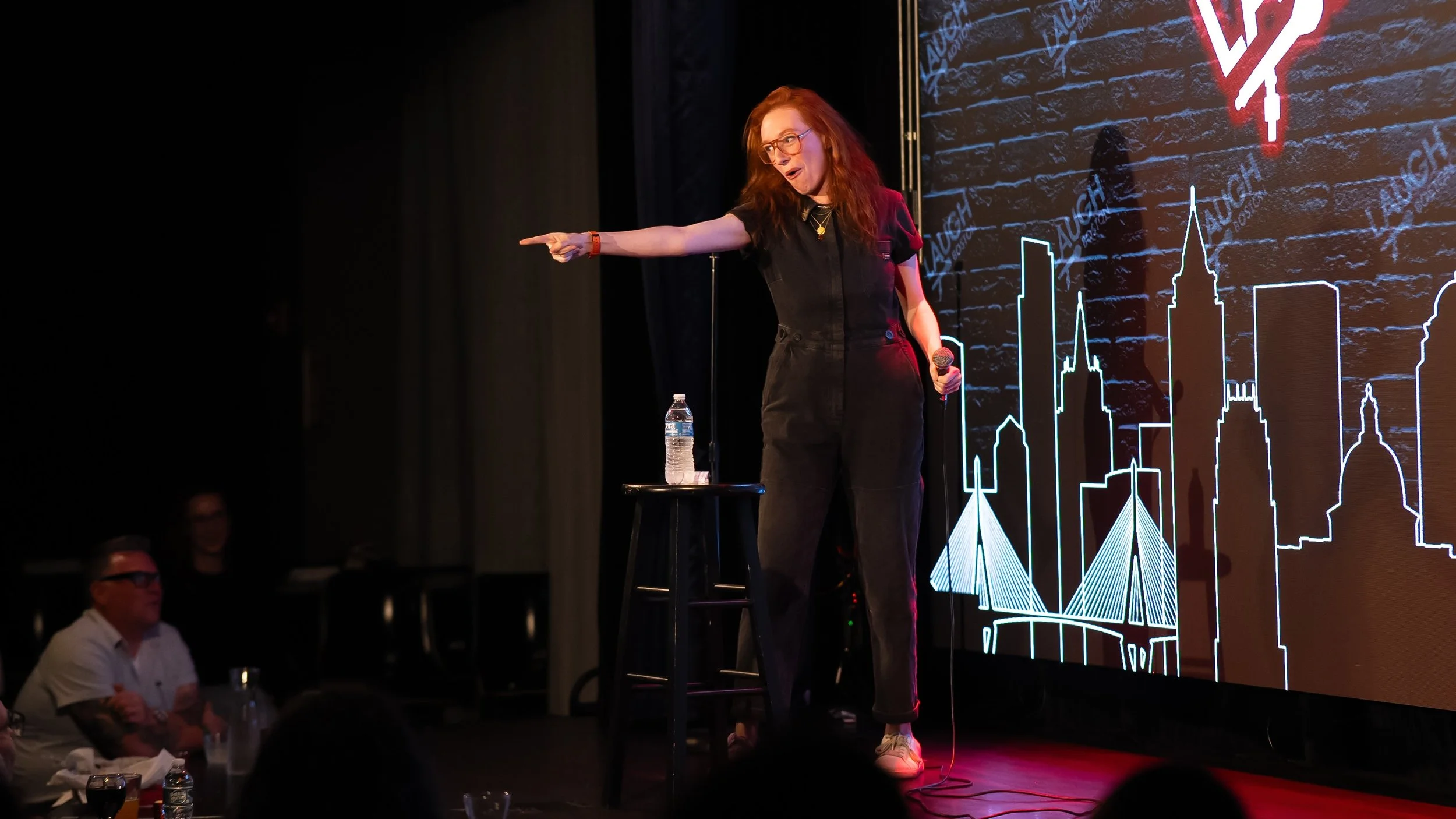 A woman with red hair, glasses, and a black jumpsuit is standing on a stage, holding a microphone in her left hand and pointing with her right hand. She appears to be performing or speaking, with an animated expression. A water bottle and a small black stool are beside her. The background features a large digital display with neon cityscape outlines and the word 'AUGUST' repeatedly written in the backdrop.
