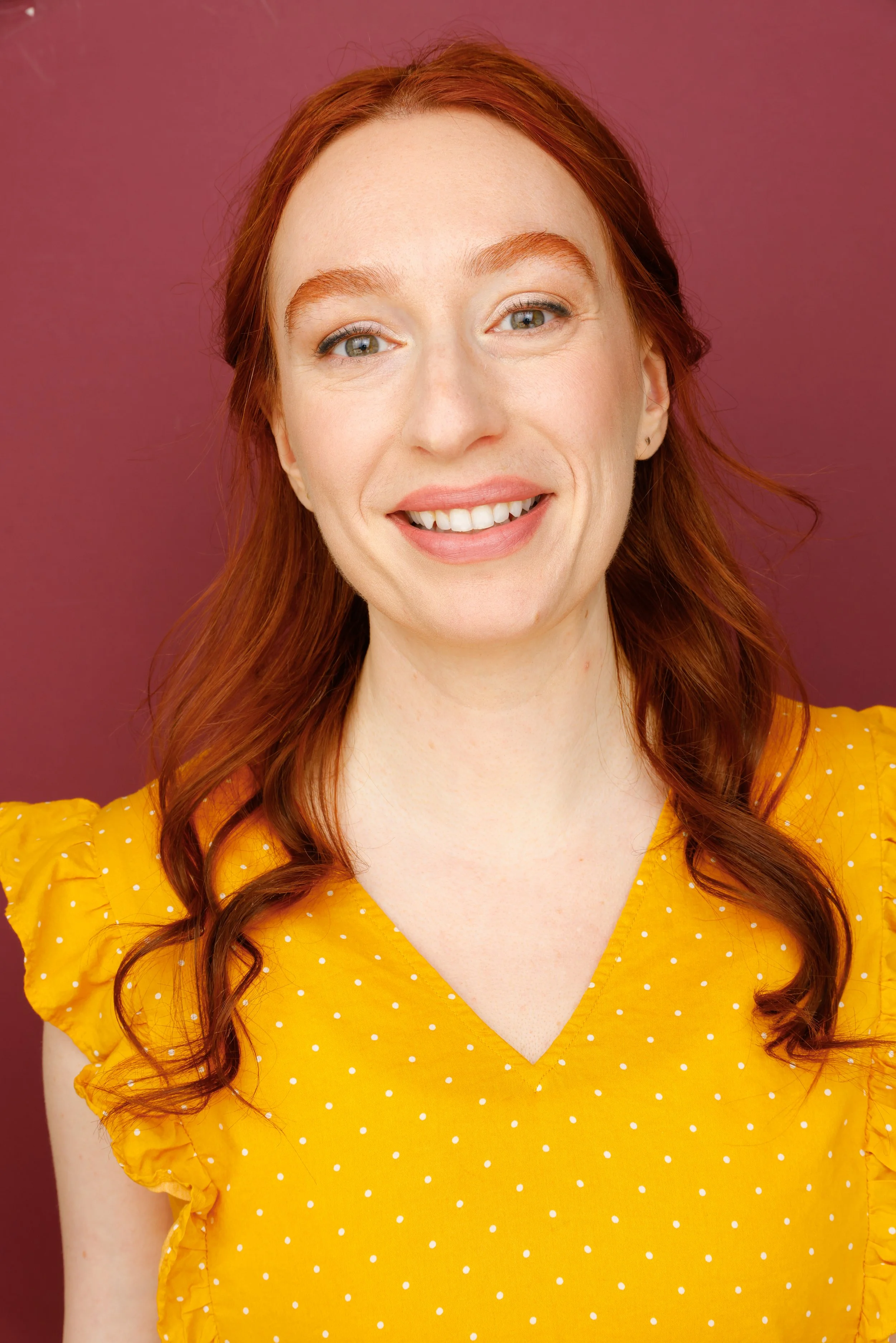 Close-up of a smiling woman with red hair wearing a yellow polka dot blouse against a mauve background.