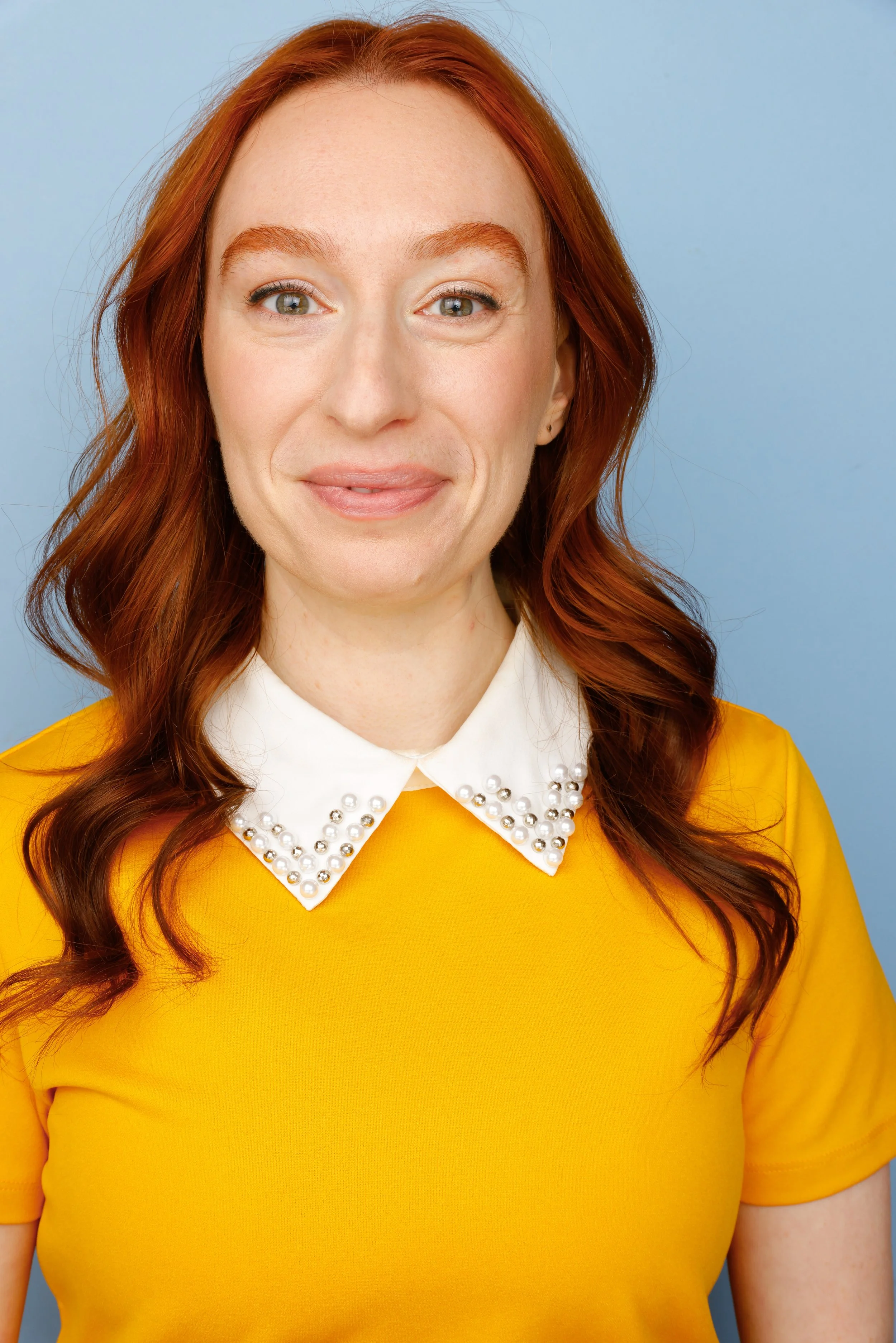 Close-up of a woman with red hair in soft curls wearing a yellow top with a white pearl-detailed collar, against a blue background.