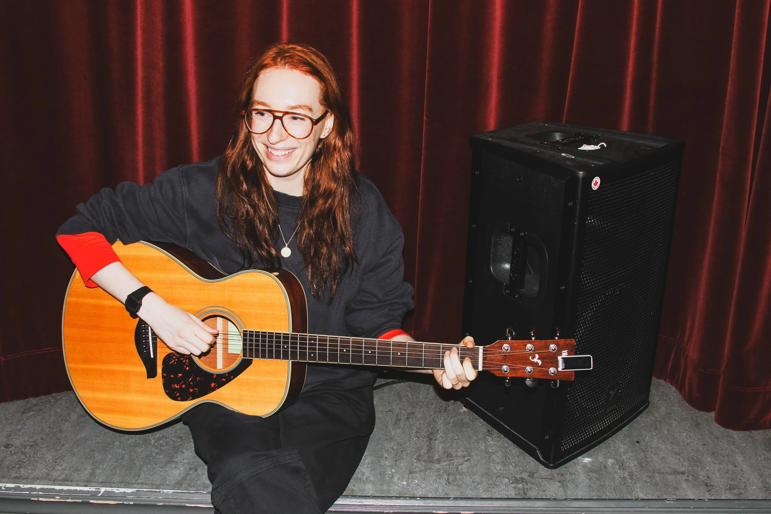 A person with long red hair, glasses, and a black hoodie playing an acoustic guitar on a stage with red curtains in the background, next to a black speaker.