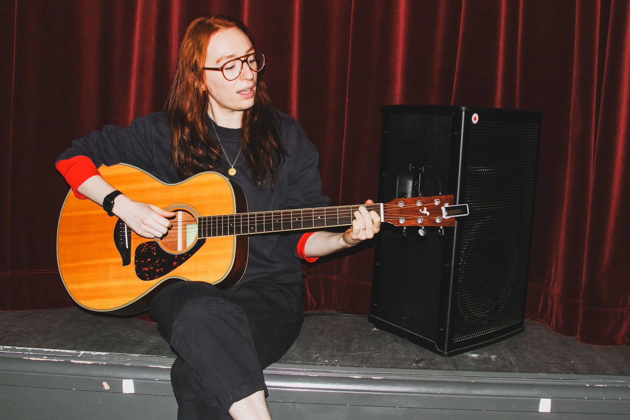 A young woman with glasses and long red hair is sitting on a stage, playing an acoustic guitar. She is wearing a black shirt with red cuffs and a necklace. Behind her is a red curtain and an amplifier.