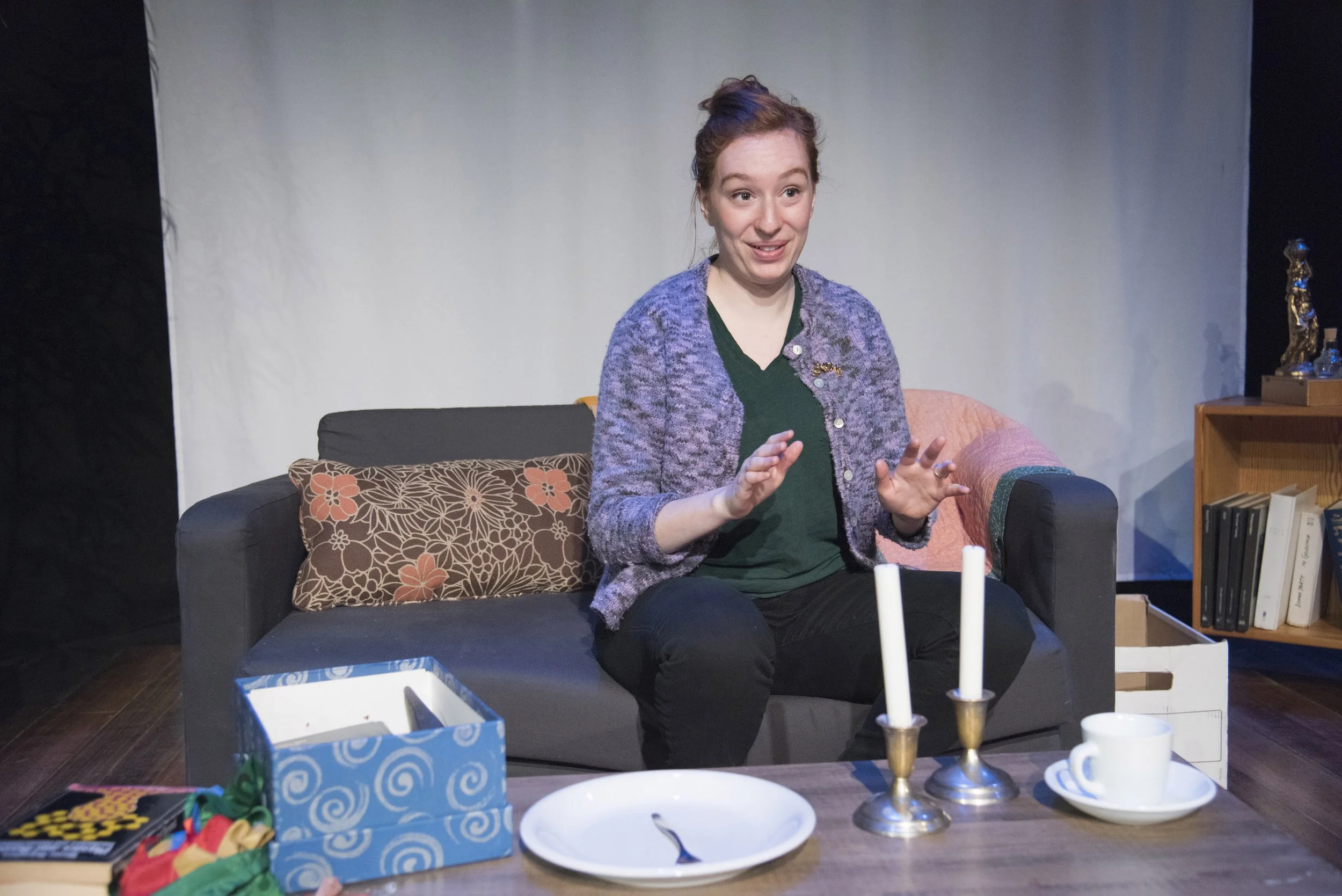 A woman with red hair, wearing a green shirt and a purple cardigan, sits on a couch in a living room, speaking with her hands. There are candles, a tissue box, and other items on the table in front of her, and a bookshelf with books and a decorative 