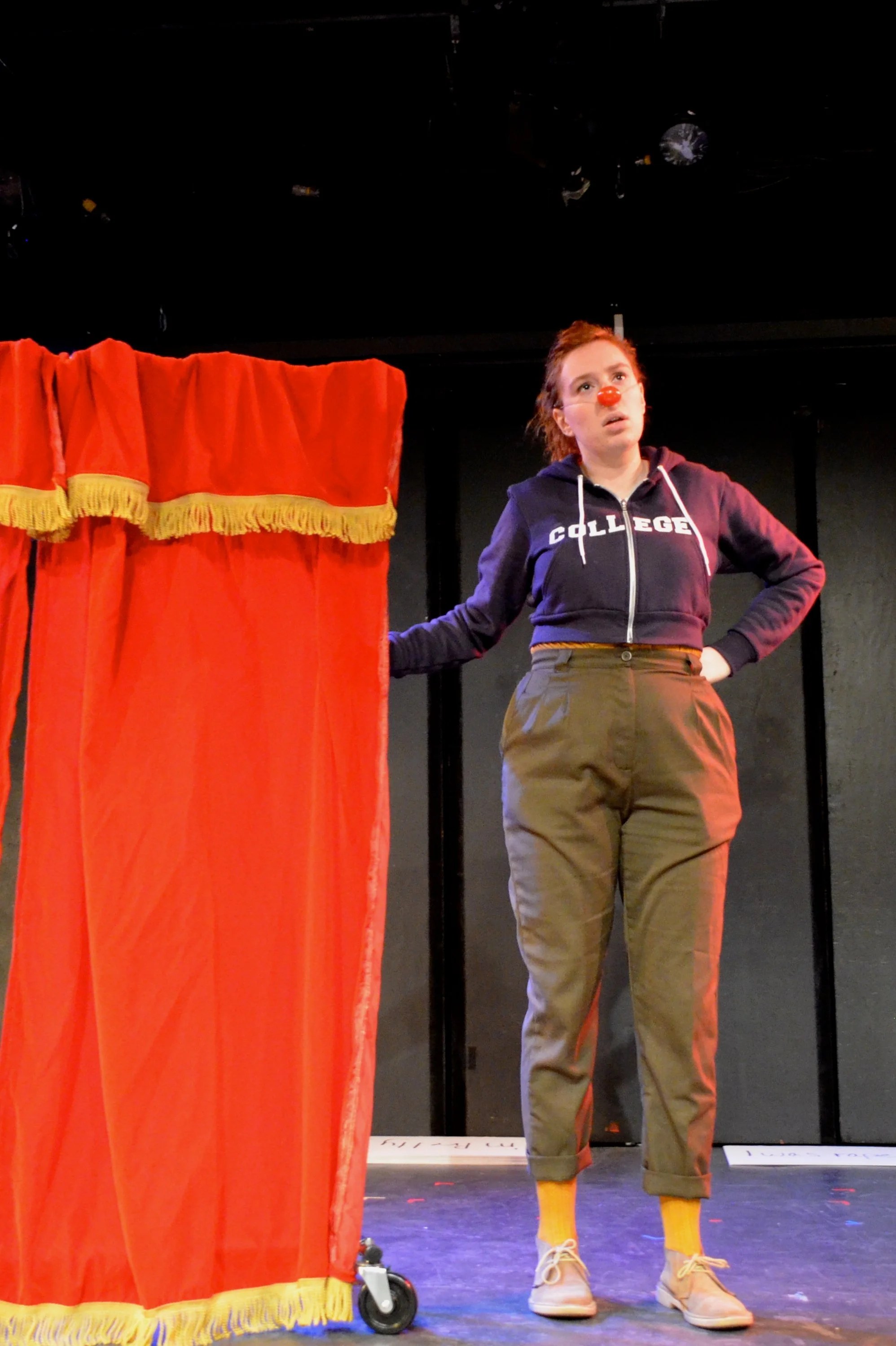 A person dressed as a clown standing on stage beside a red curtain, wearing a navy hoodie with the word 'college' on it, khaki pants, yellow socks, and beige sneakers, with a red clown nose and a surprised expression.