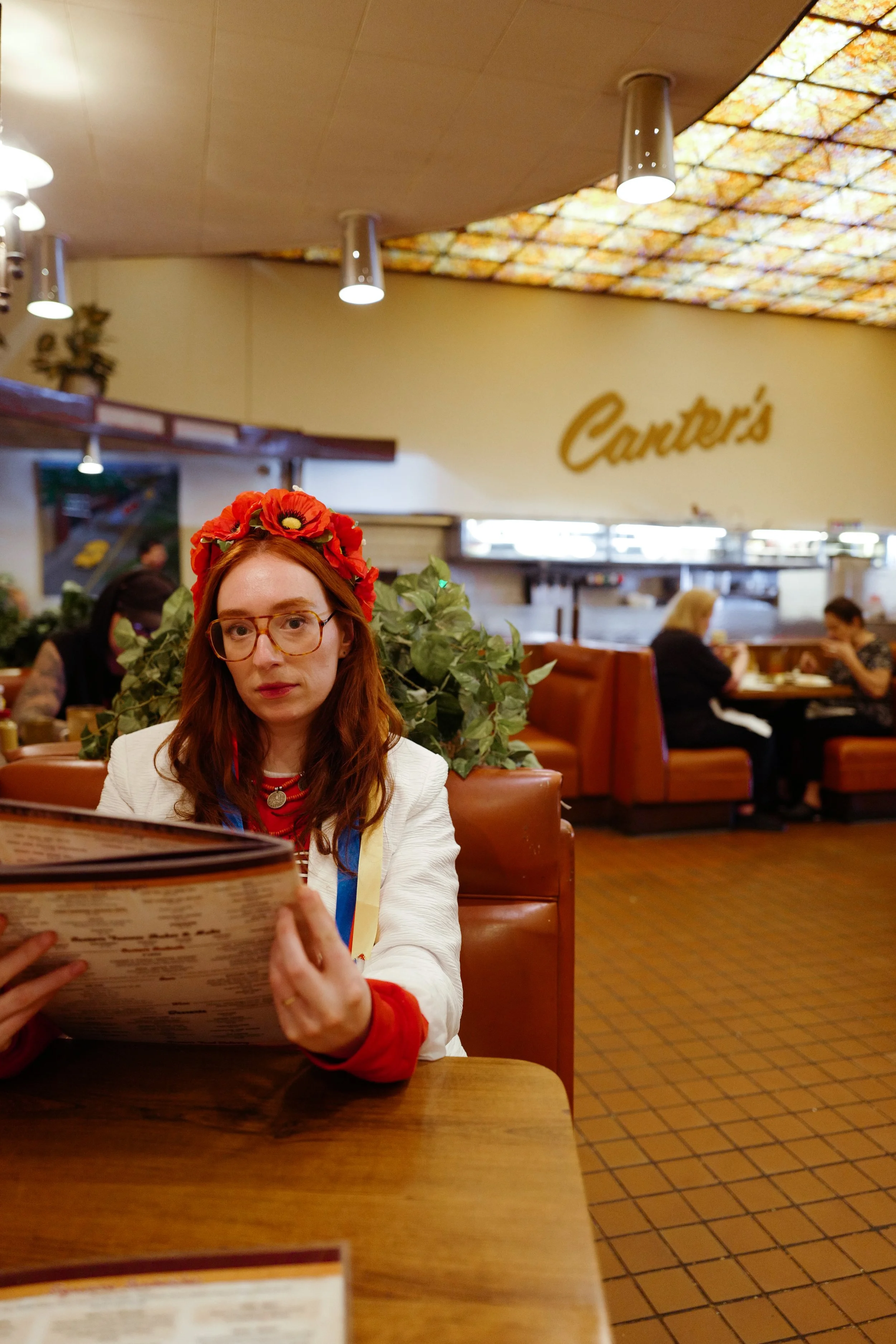 A woman with red hair, glasses, a floral headband, and a white blazer with red wristbands is sitting at a restaurant table holding a menu. In the background, there are other people dining and a sign that reads 'Cincinnati' on the wall.