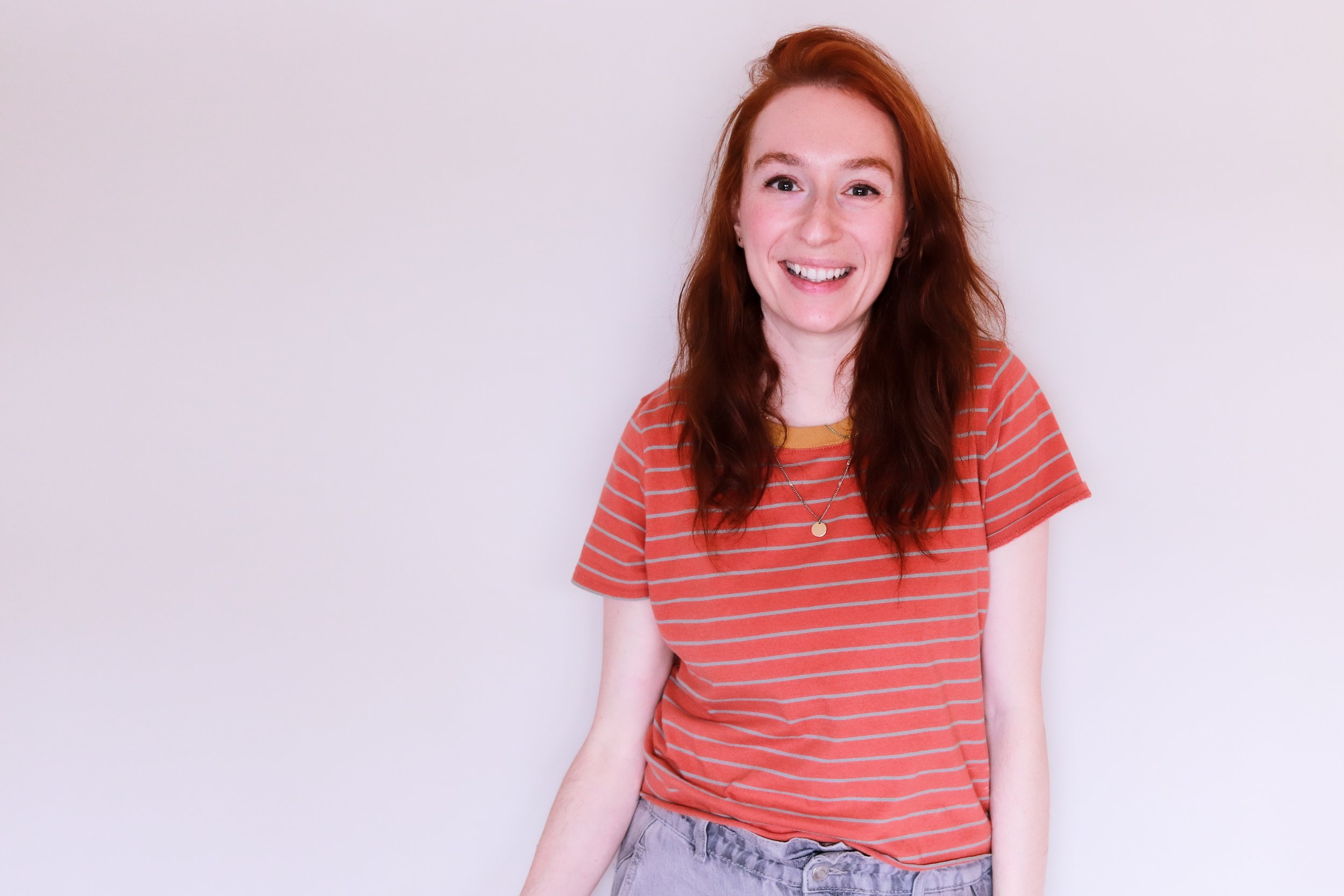 Young woman with long red hair smiling, wearing a red striped T-shirt and light gray shorts, standing against a plain white wall.