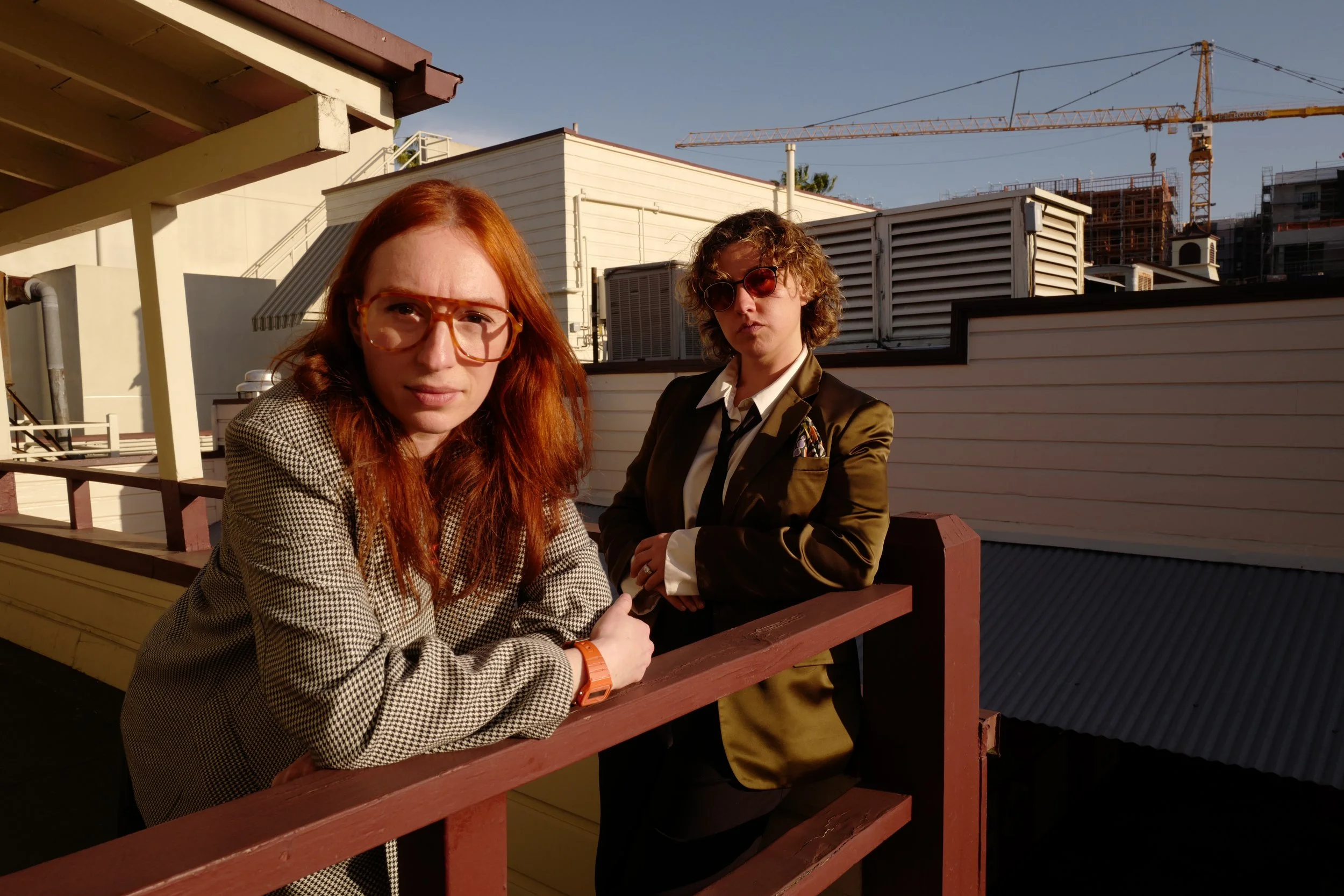 Two women with red and curly hair and sunglasses, dressed in business attire, leaning on a railing outside on a rooftop patio with buildings, a crane, and a clear blue sky in the background.