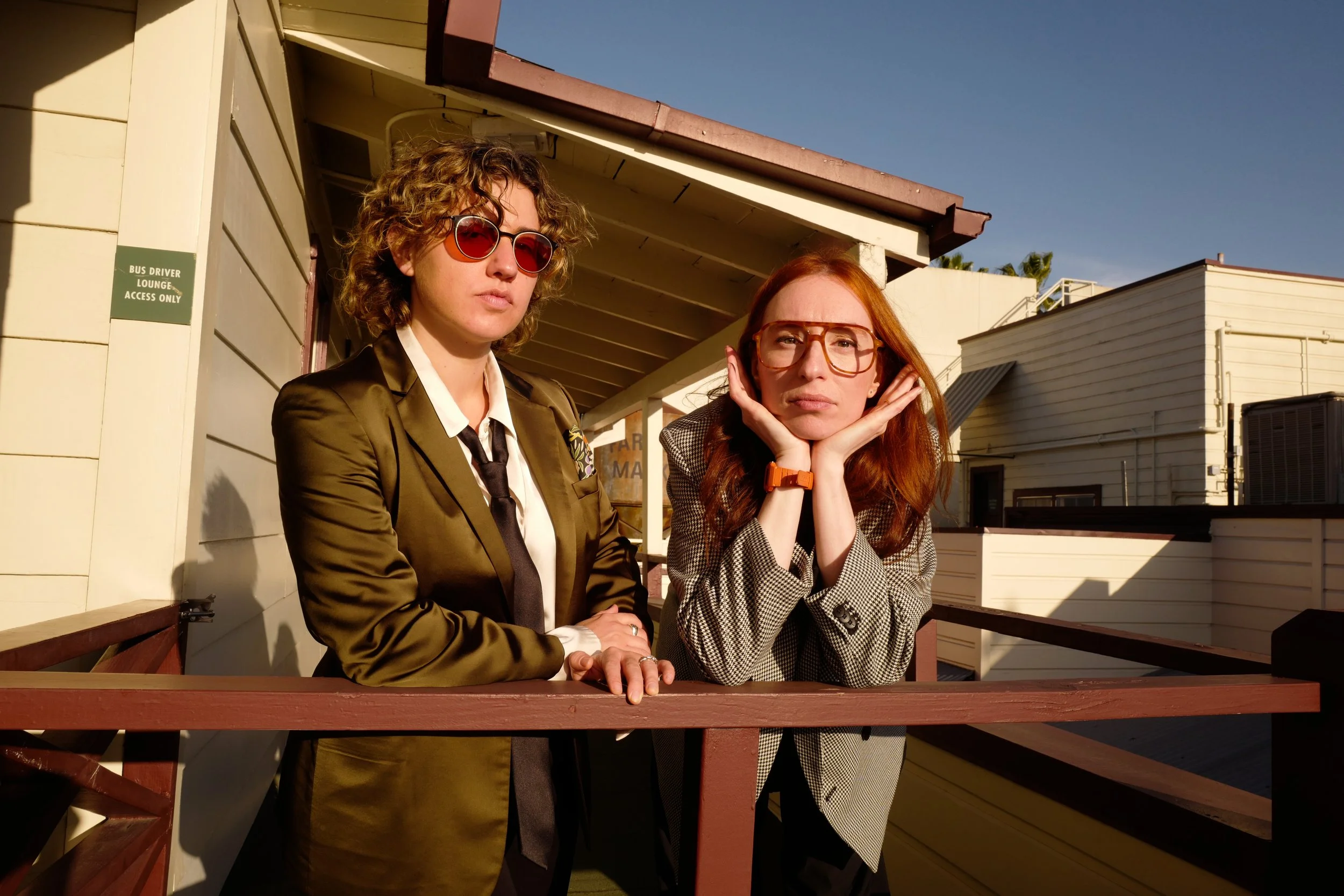 Two women with curly hair and glasses sitting on a balcony under clear blue sky, one with red hair, the other with brown hair, wearing stylish clothing.