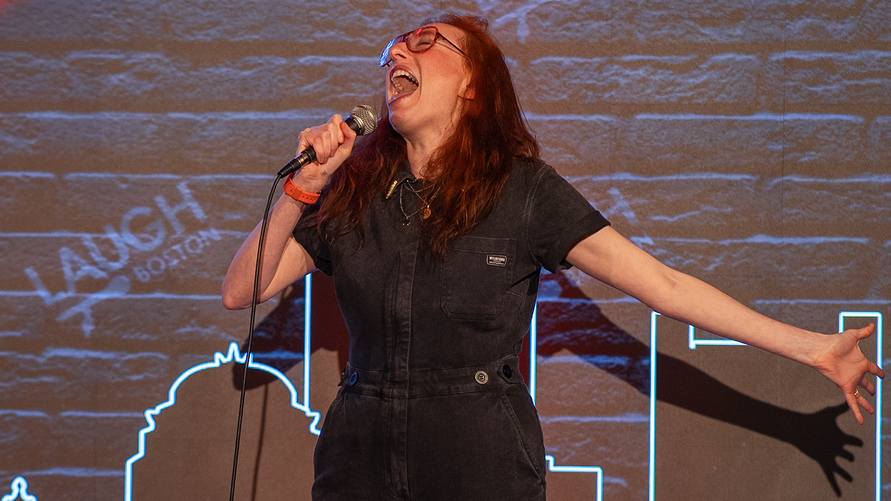 A woman with red hair and glasses singing passionately into a microphone with her arms outstretched on stage.