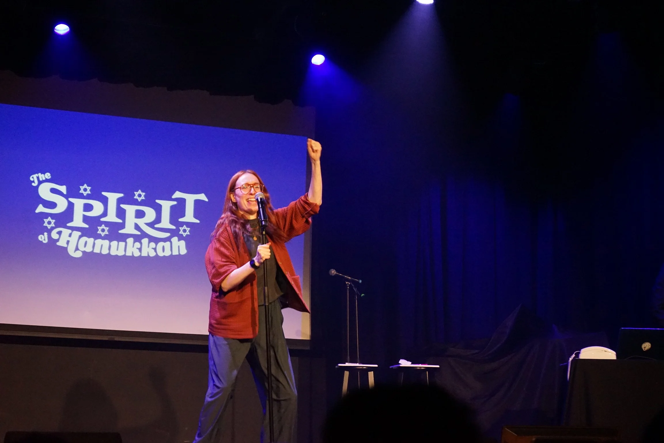 A woman wearing glasses and a red checkered jacket speaks into a microphone on stage, raising her right fist. There is a large screen behind her with the text "The Spirit of Hanukkah" and Star of David symbols. The stage is dark with blue lighting.