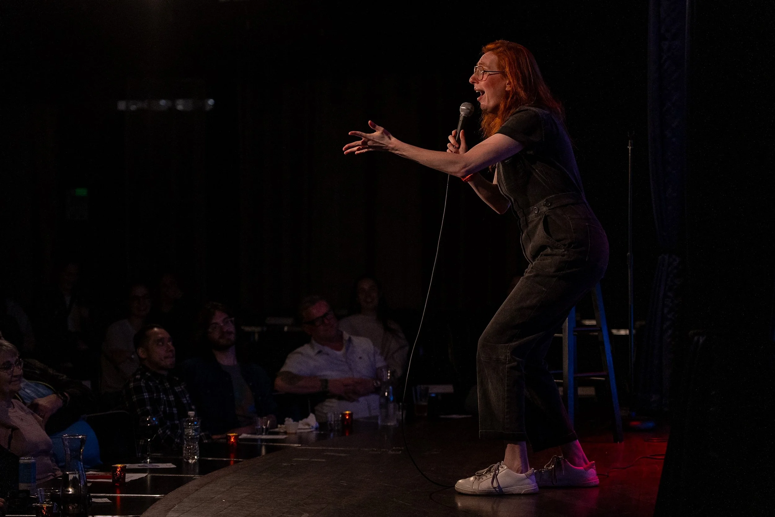 A woman with glasses and red hair performing stand-up comedy on stage, holding a microphone, in front of an audience.