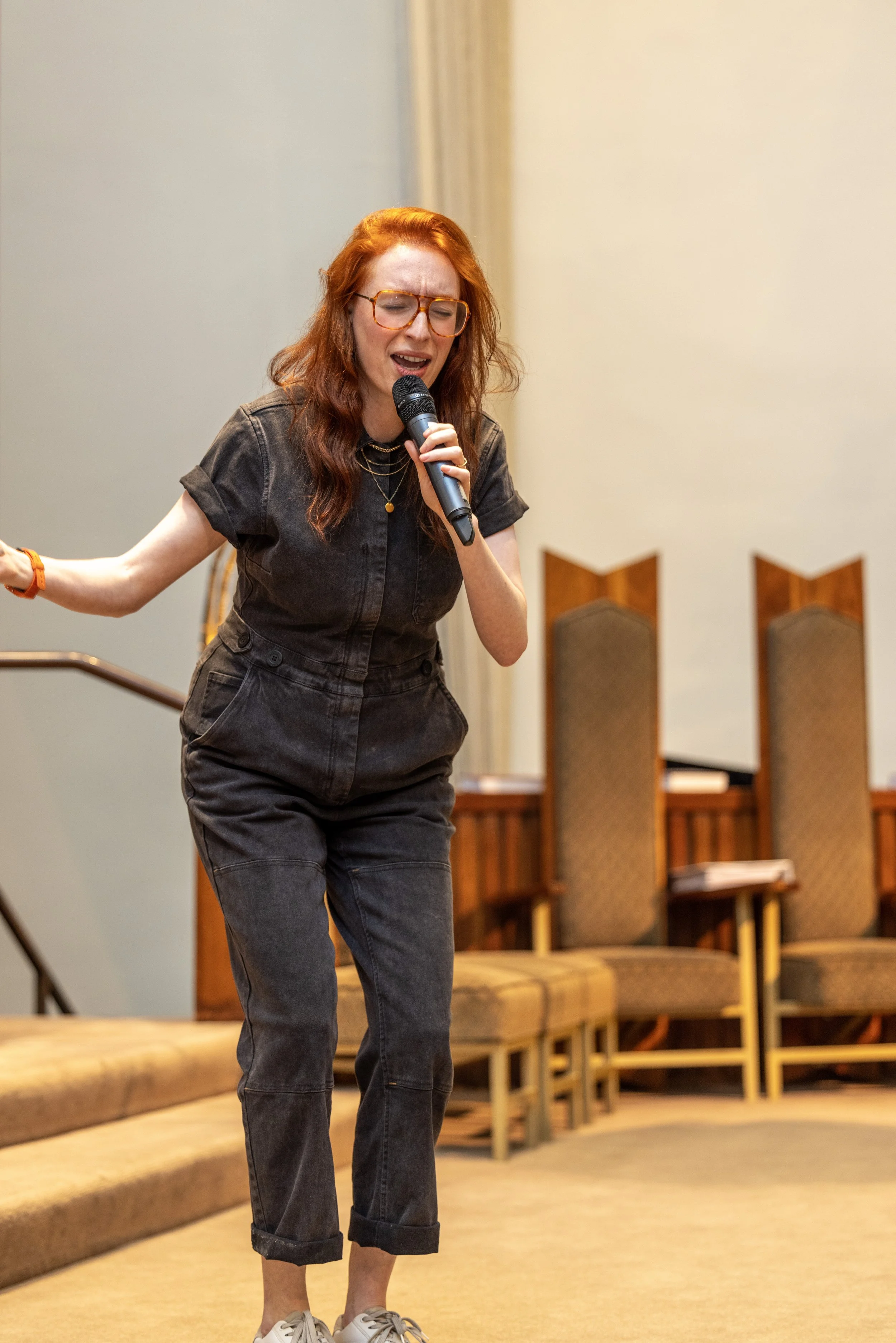 A woman with red hair and glasses singing passionately into a microphone in a room with beige chairs and carpeted floor.