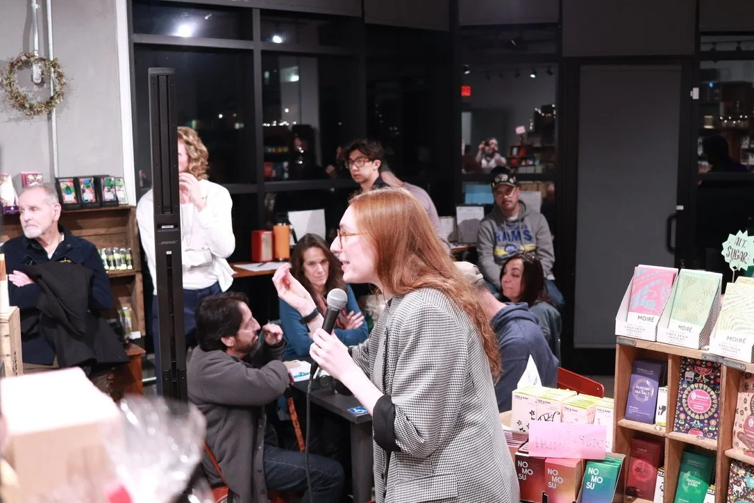 A woman with red hair and glasses holding a microphone speaking to a small crowd at a bookstore event, with books and shelves visible in the background.