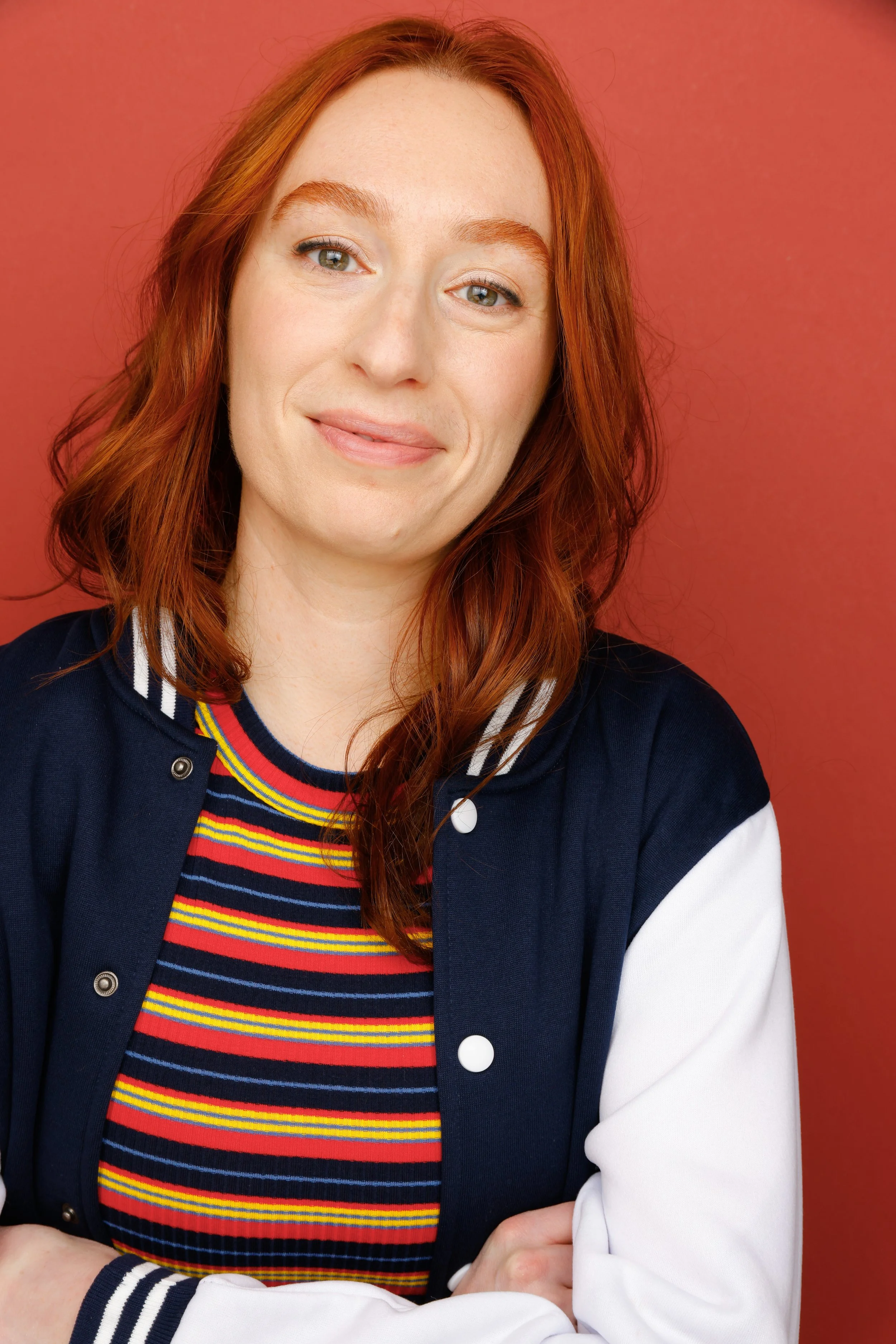 A portrait of a woman with red hair and blue eyes, wearing a navy blue jacket with white and colorful striped shirt underneath, with a red background.