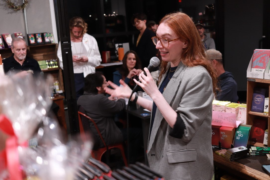 A woman with long red hair and glasses speaking into a microphone at a book store event, with a group of people seated and standing around her.