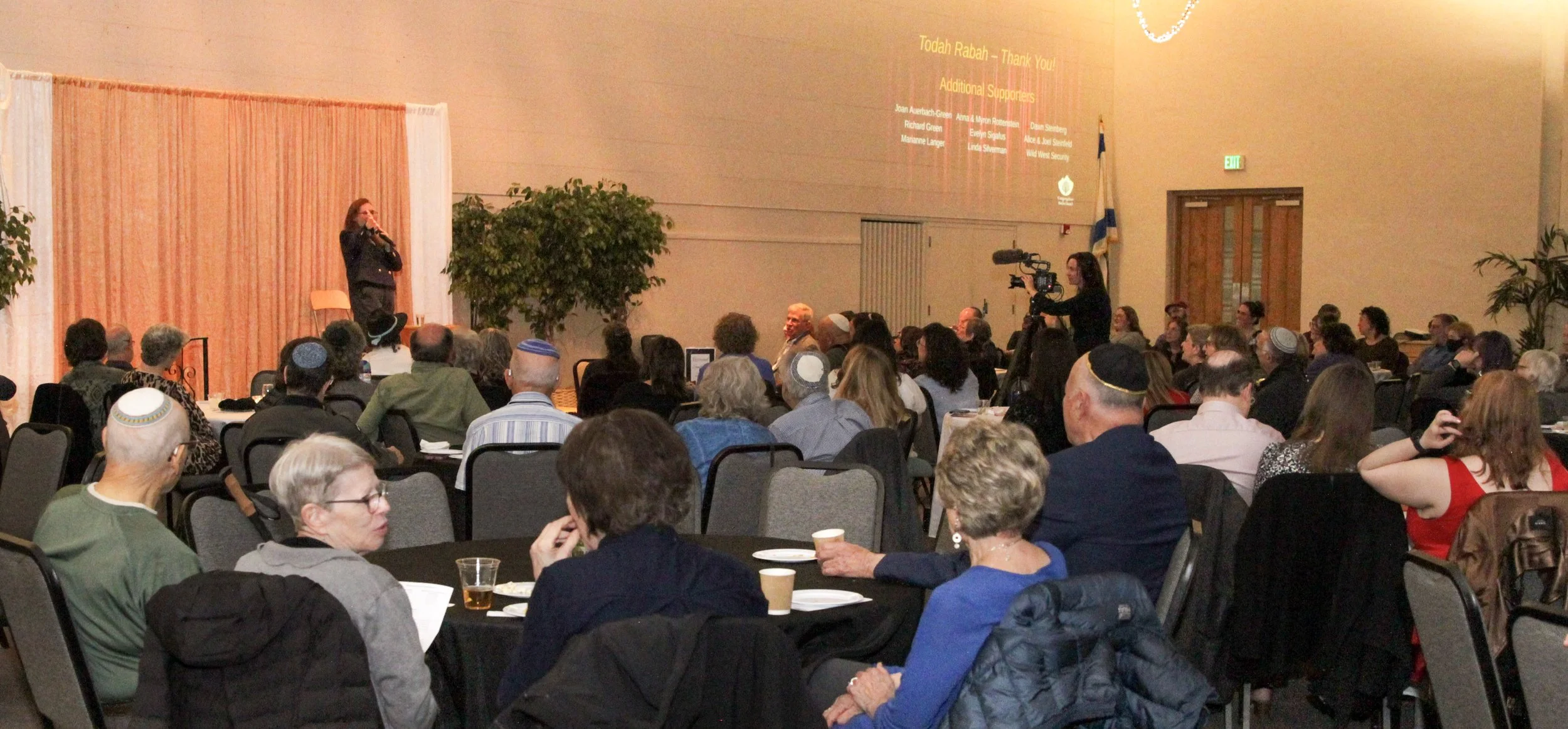 A speaker on stage addressing an audience in a large room, with a projector screen displaying the event title and sponsors, and a video camera operator recording the event.