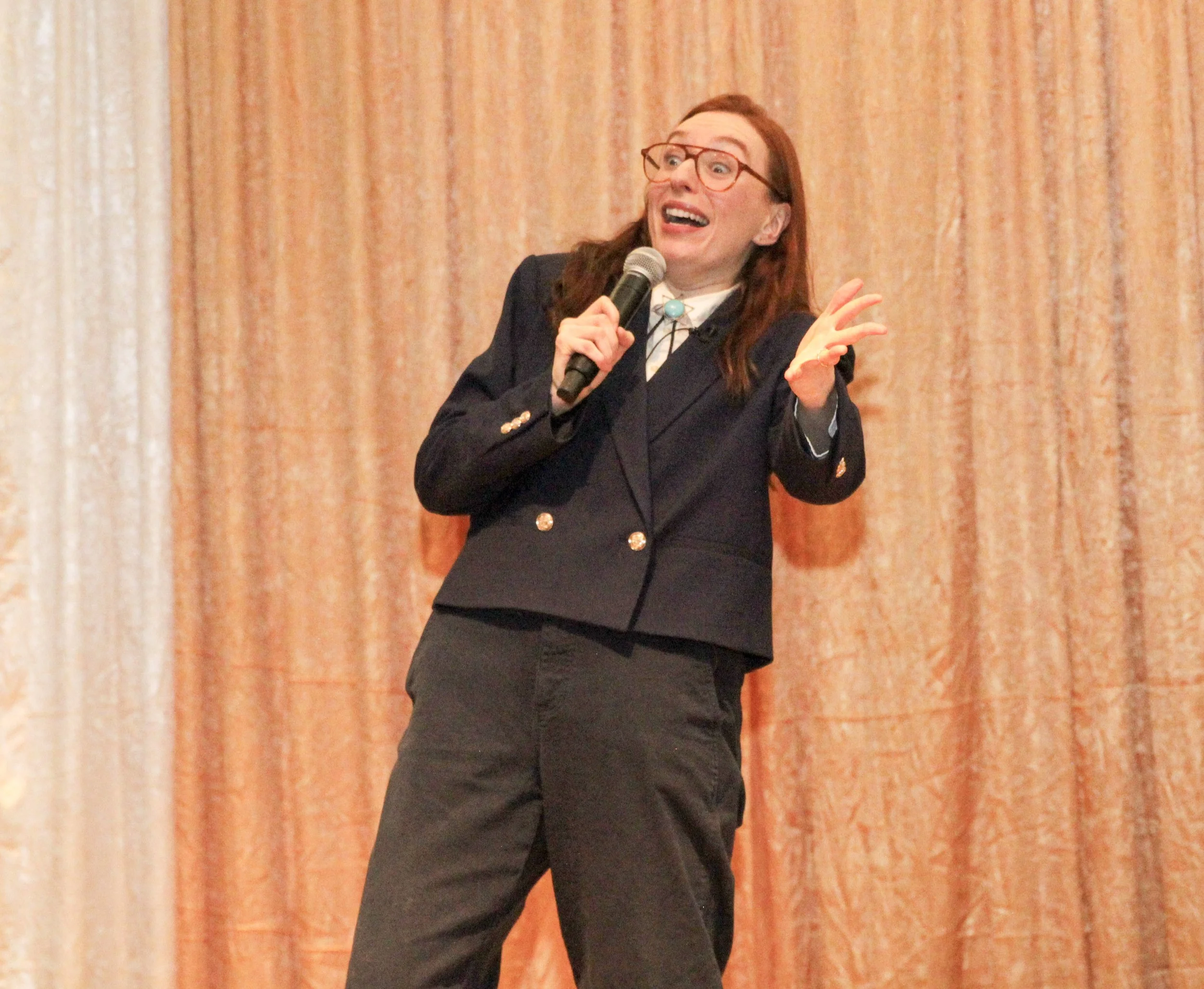 A woman with red hair and glasses wearing a black blazer and pants, holding a microphone, standing in front of a curtain.