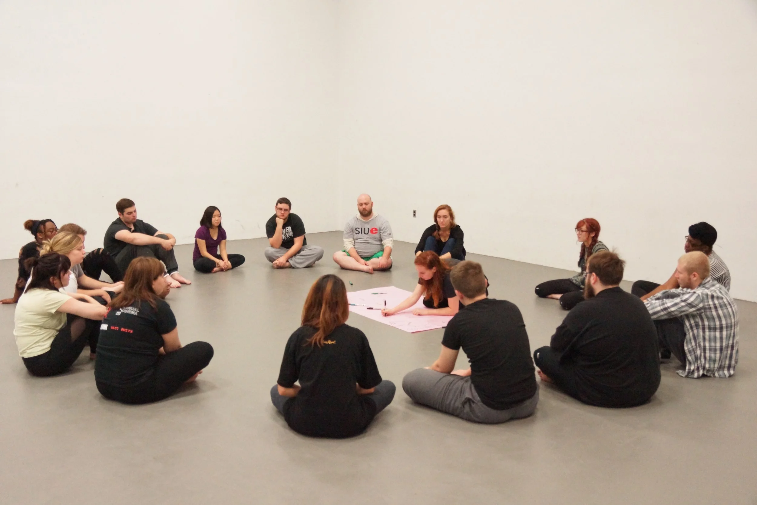 Group of people sitting in a circle on the floor inside a plain white room, engaging in a discussion or activity.