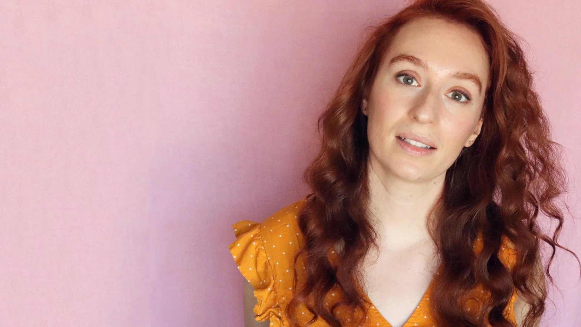 Portrait of a young woman with curly red hair wearing a mustard yellow dress with small white polka dots, standing against a pink wall.