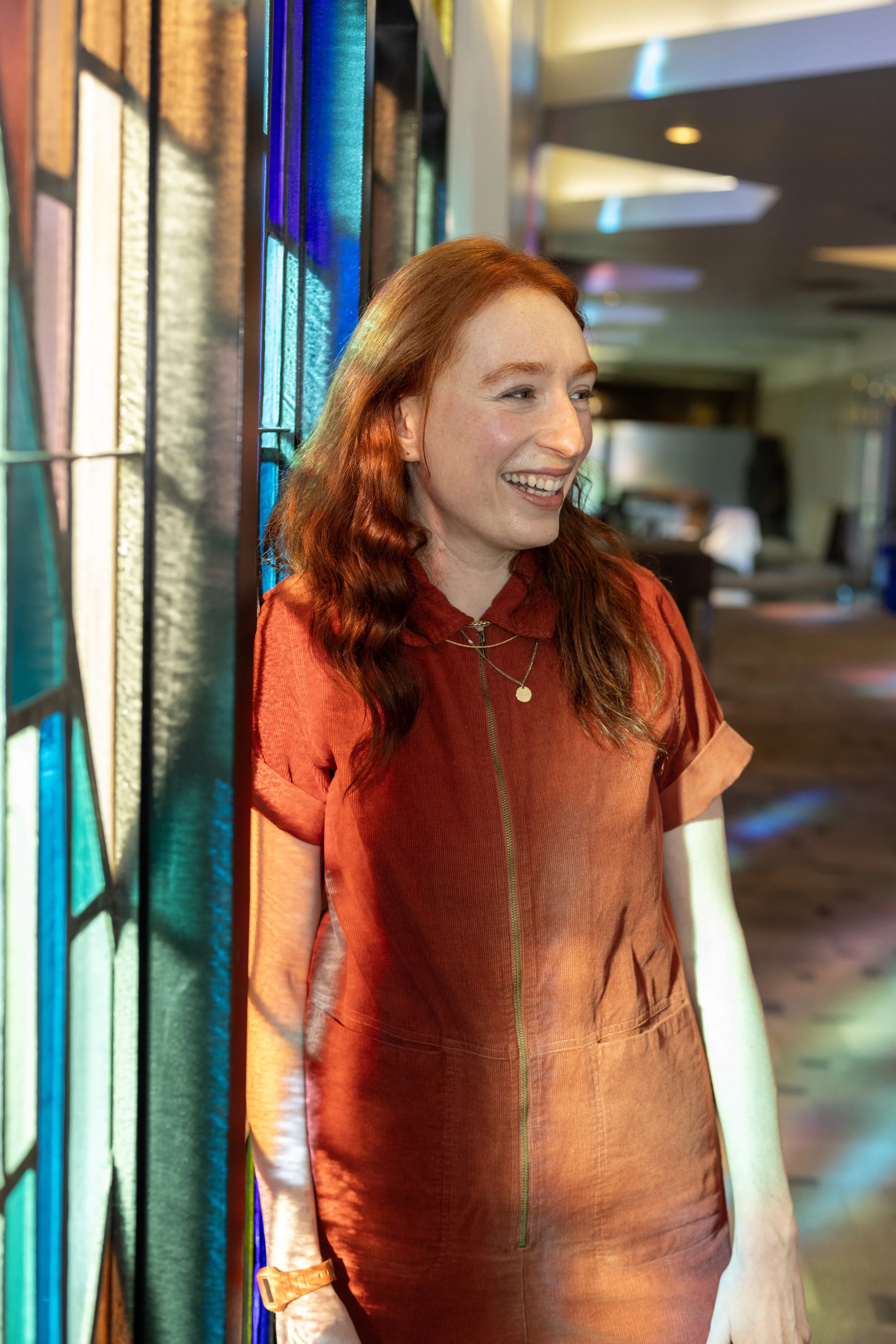 A woman with red hair smiling, standing next to stained glass windows with colorful light reflecting off them, inside a modern building.