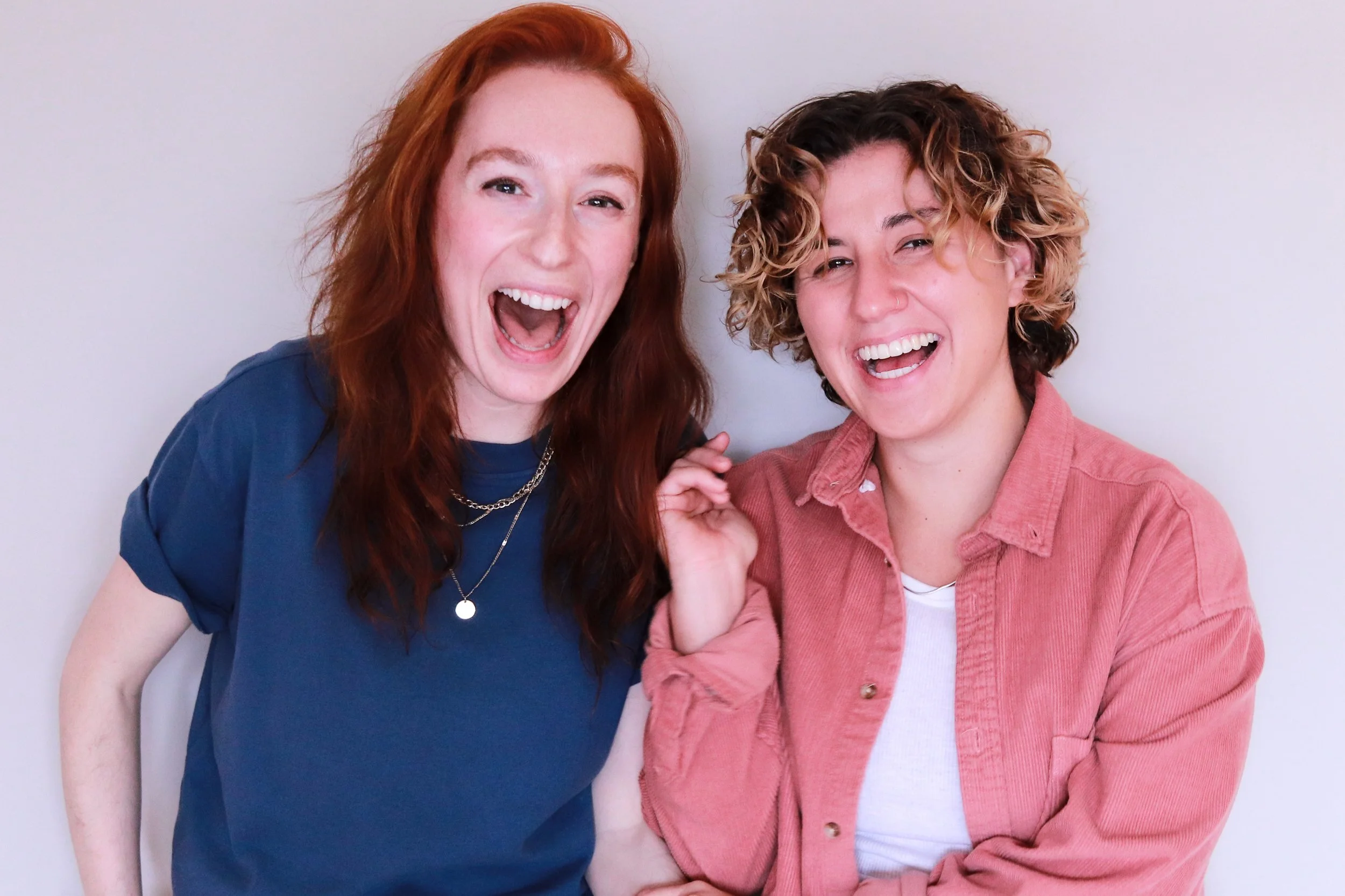 Two women smiling and laughing together, one with red hair and the other with curly light brown hair, standing against a plain white background.