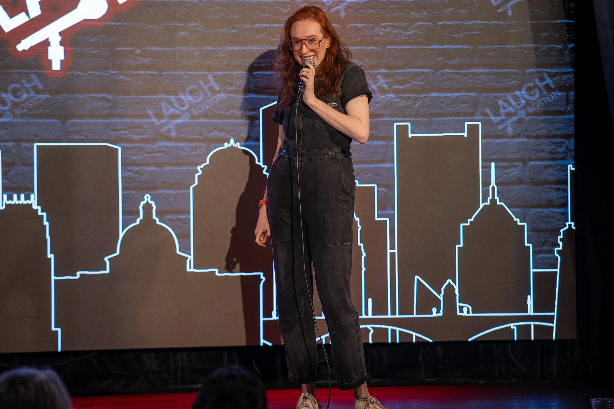 A woman with red hair, glasses, and a black jumpsuit holding a microphone on stage with a city skyline outline in neon behind her.